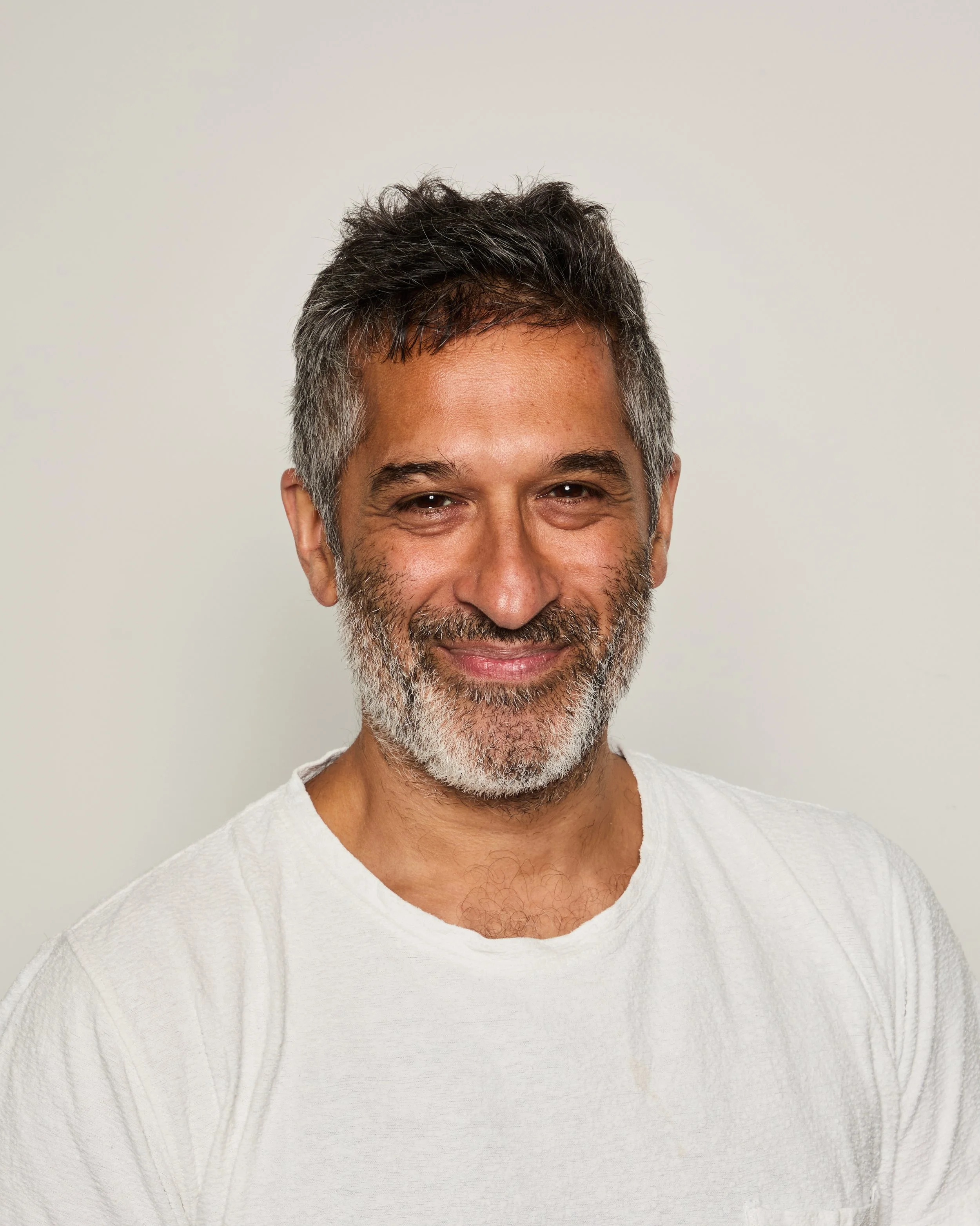 Close-up of a smiling middle-aged man with short salt-and-pepper hair, a beard, wearing a white textured shirt, standing against a plain light background.