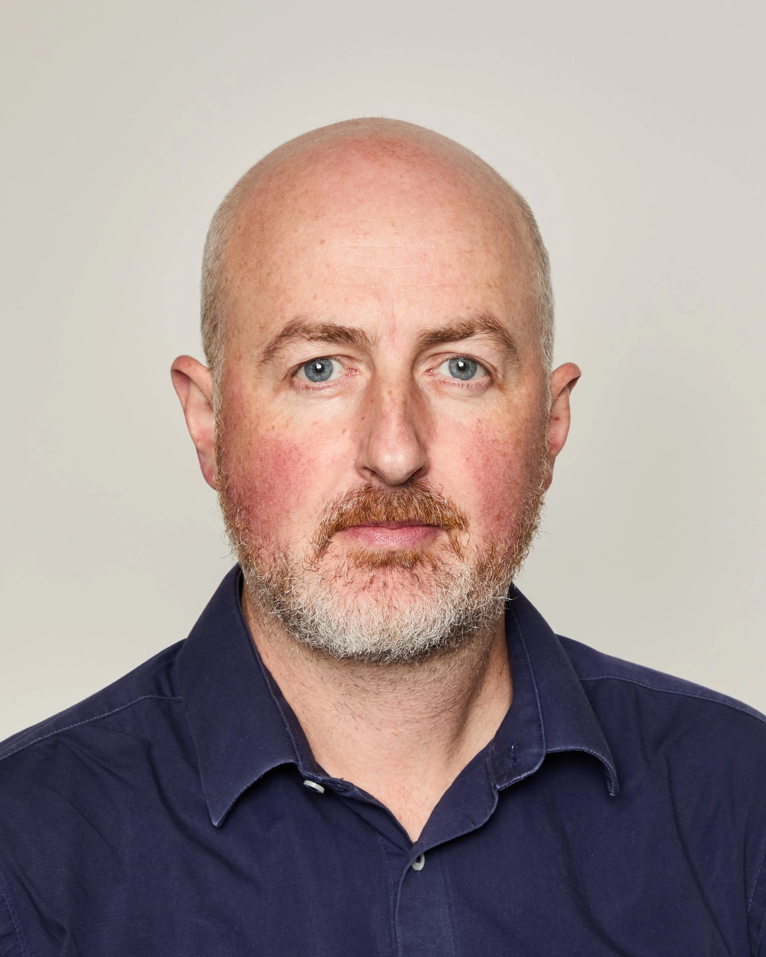 Close-up of a middle-aged man with light skin, blue eyes, a reddish beard, and a bald head, wearing a dark blue shirt.