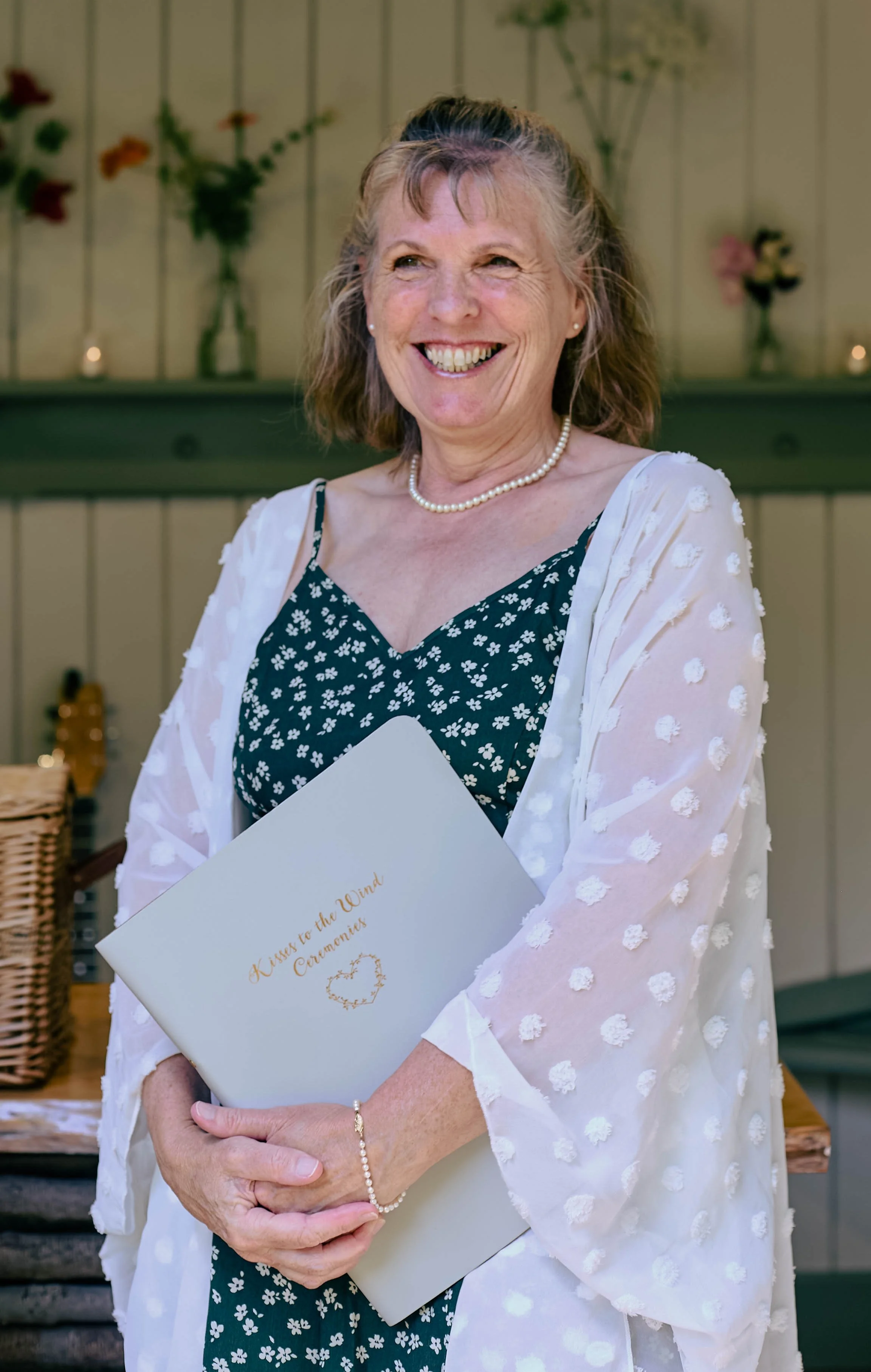 A woman smiling while holding a white  that reads 'Kisses to the Wind' with a heart. She is wearing a dress with white floral pattern, a pearl necklace, and a white cardigan with pom-pom detailing. She is standing indoors with flowers and candles in 