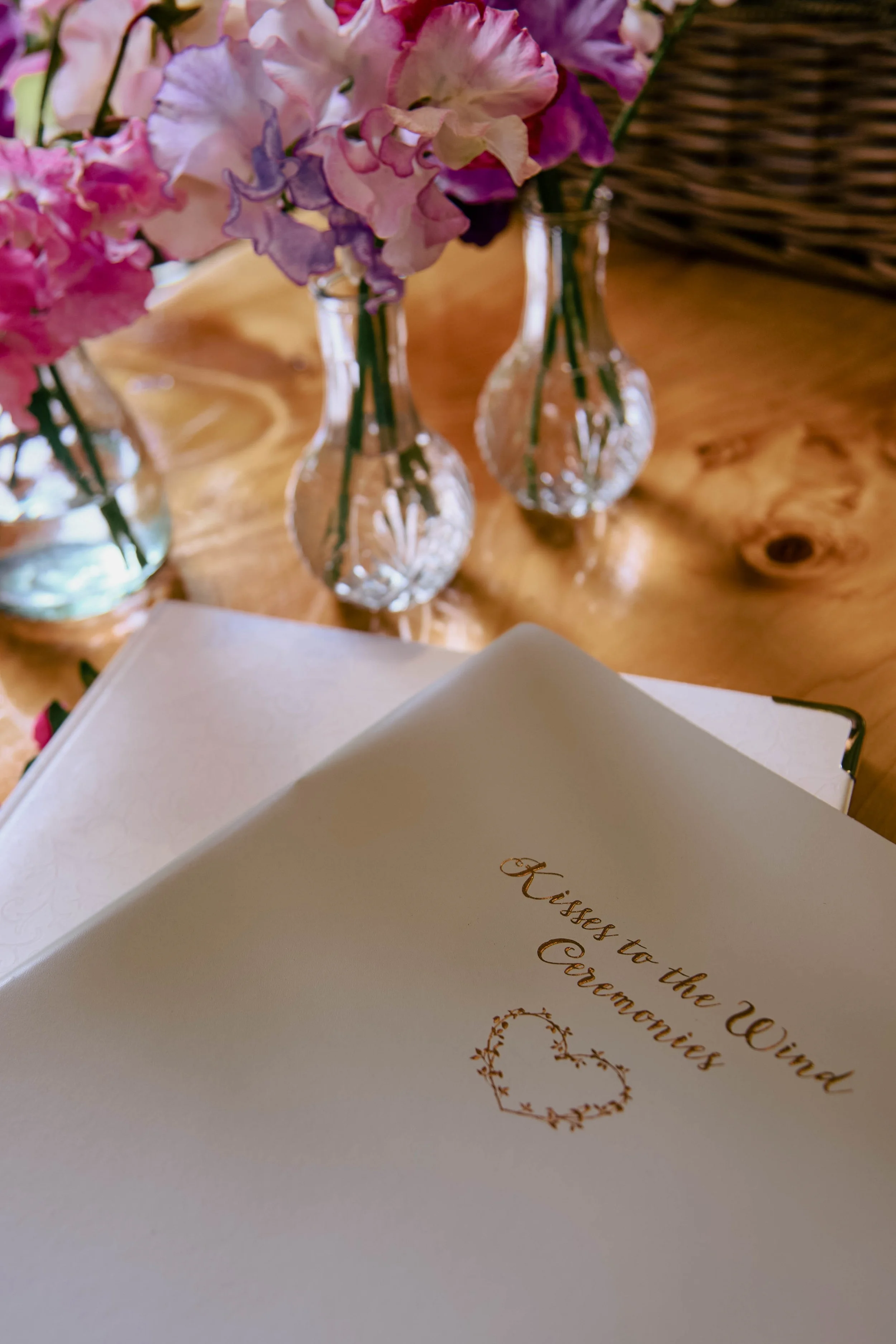 Close-up of a white book with gold text that reads "Kisses to the Wind Ceremonies". Behind the book are three glass vases with pink, purple, and white hydrangea flowers, set on a wooden surface.