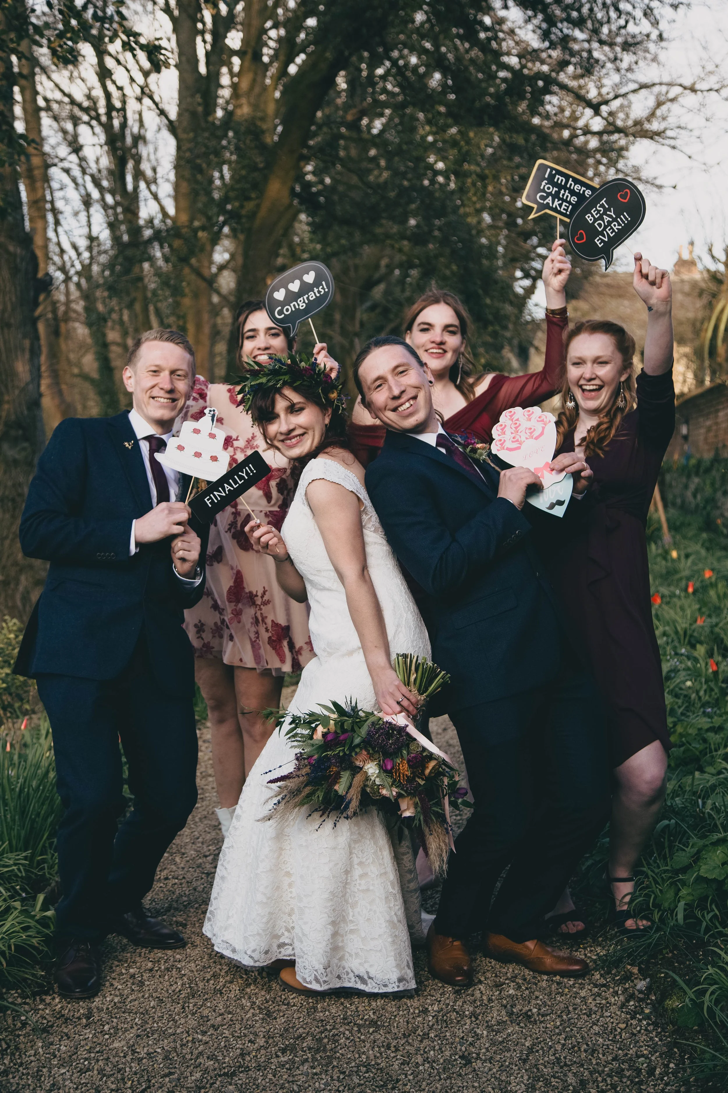 A group of six people celebrating a wedding outdoors, with the bride in a white dress holding a bouquet and smiling, surrounded by friends holding congratulatory and celebration signs, with trees and a garden path in the background.