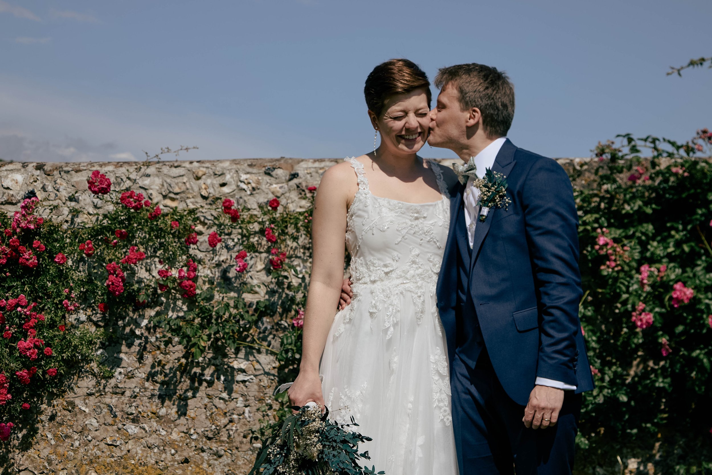 A bride and groom share a kiss outdoors against a stone wall with pink flowers, under a blue sky.