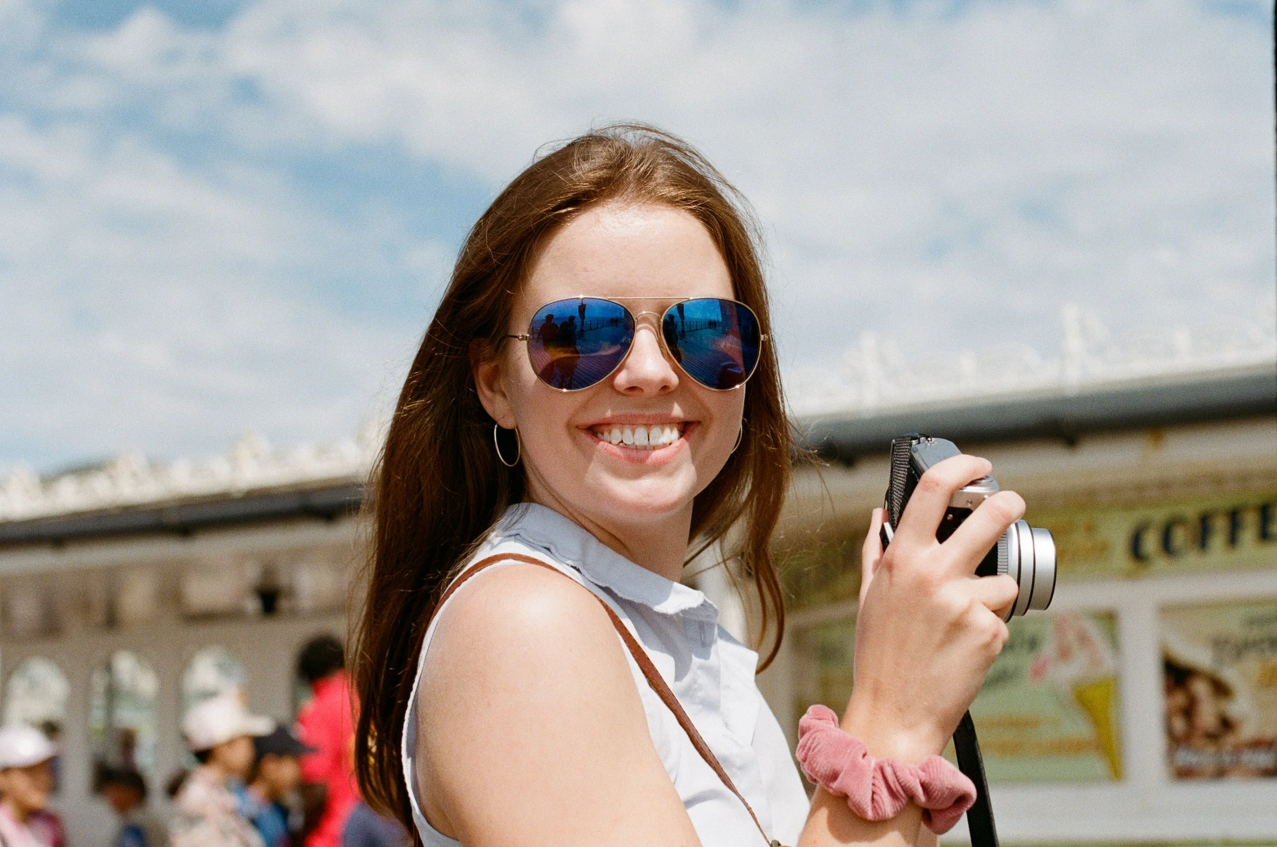 A woman with long brown hair, wearing sunglasses, smiling and holding a camera, standing outdoors on a sunny day with a blue sky and a building with a yellow sign in the background.