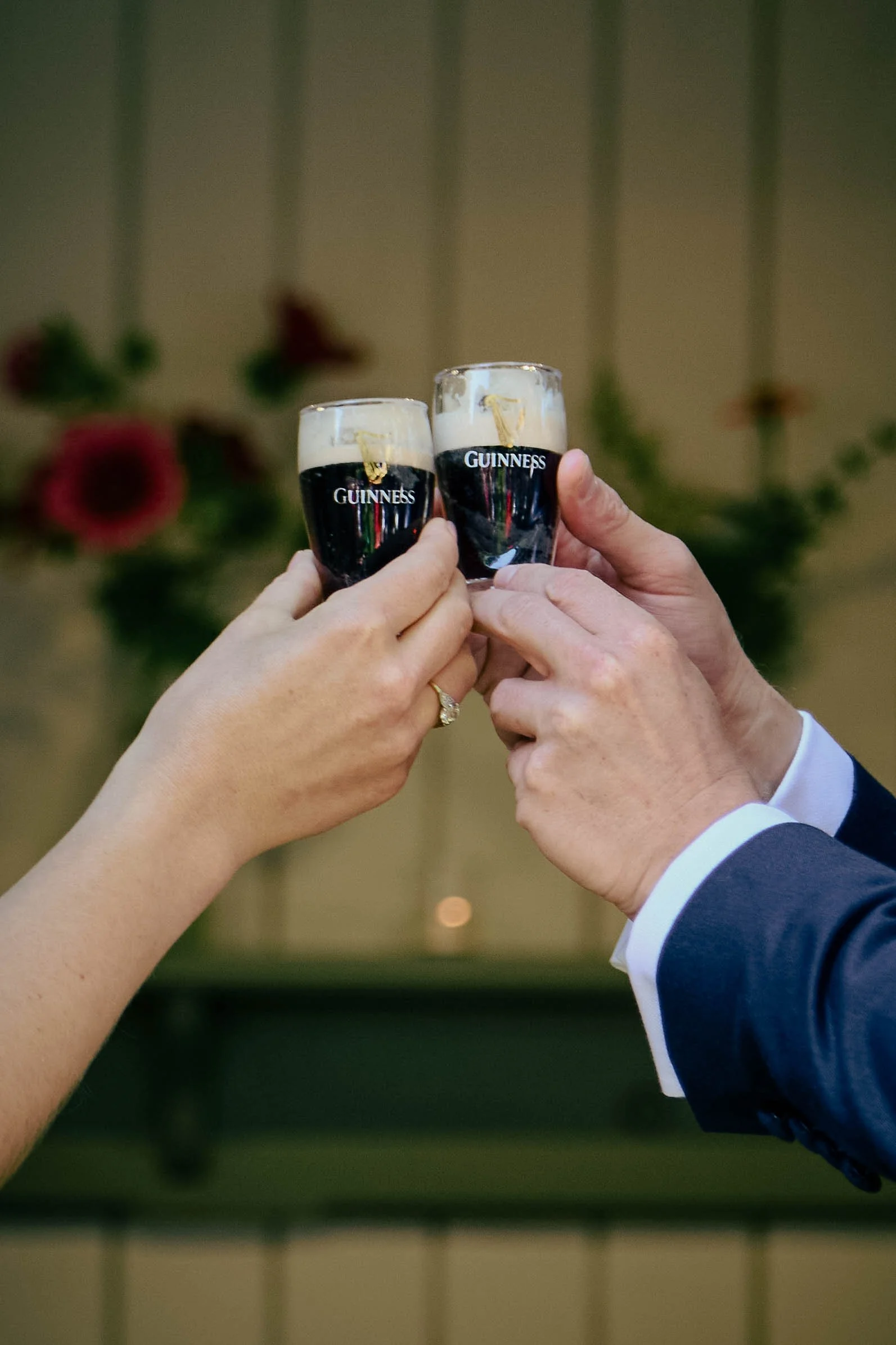 Two people clinking mini glasses of Guinness beer in a celebration, one person wearing a wedding ring and the other in formal attire.