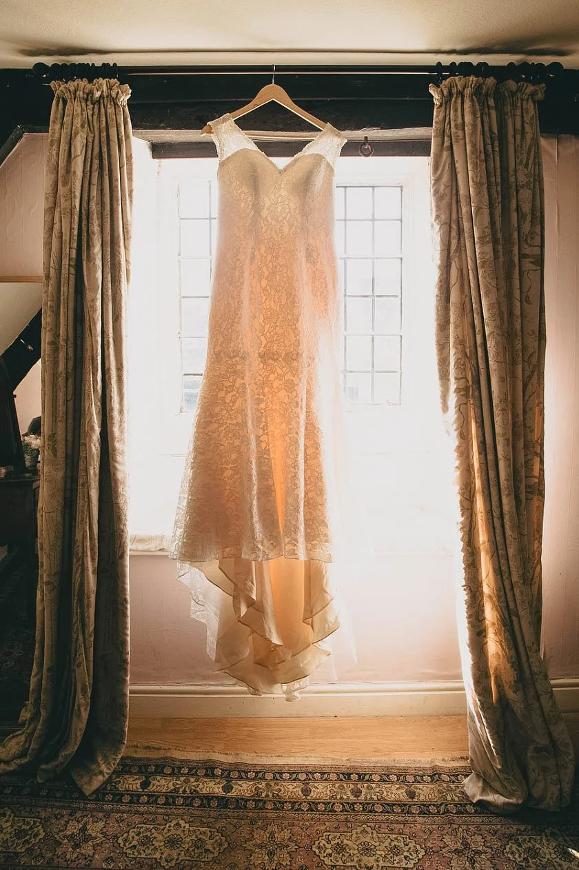 Wedding dress hanging in front of a window with curtains on either side.
