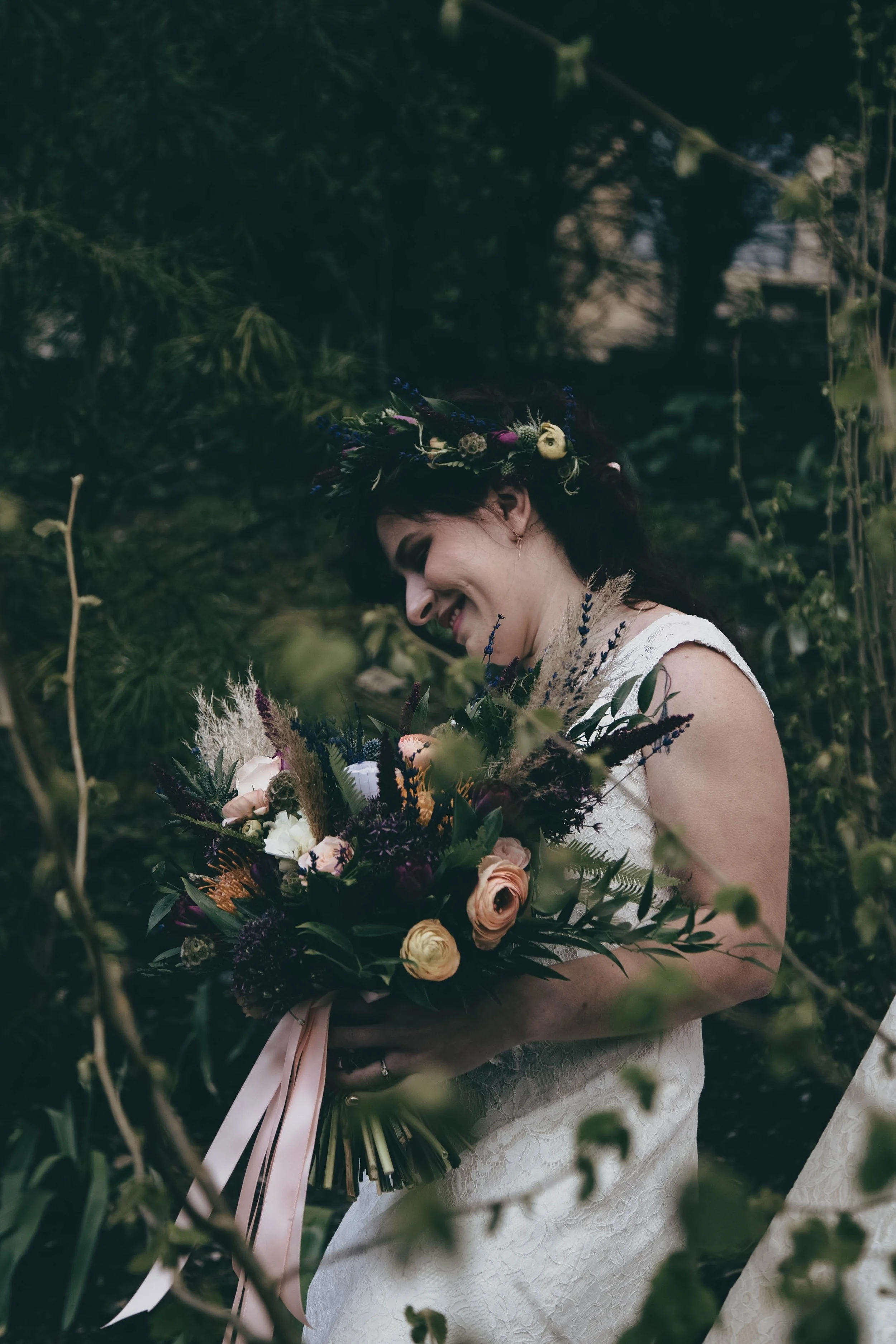 A woman wearing a white lace dress and a floral crown is holding a bouquet of flowers in an outdoor natural setting, partially obscured by greenery.
