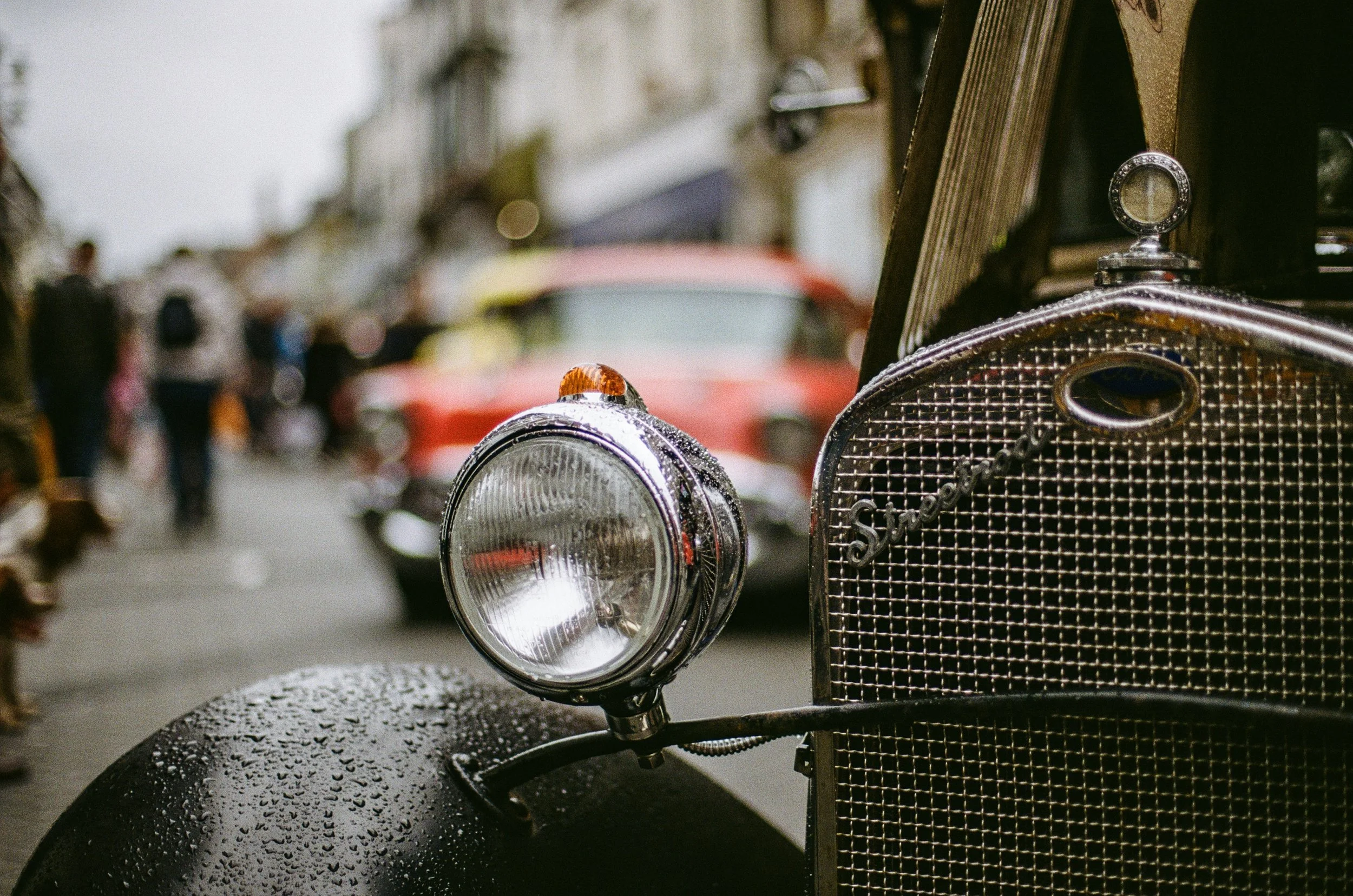 Close-up of a vintage black car with raindrops on the hood, showing the front grille, headlight, and a side mirror, with a blurred background of a street scene.