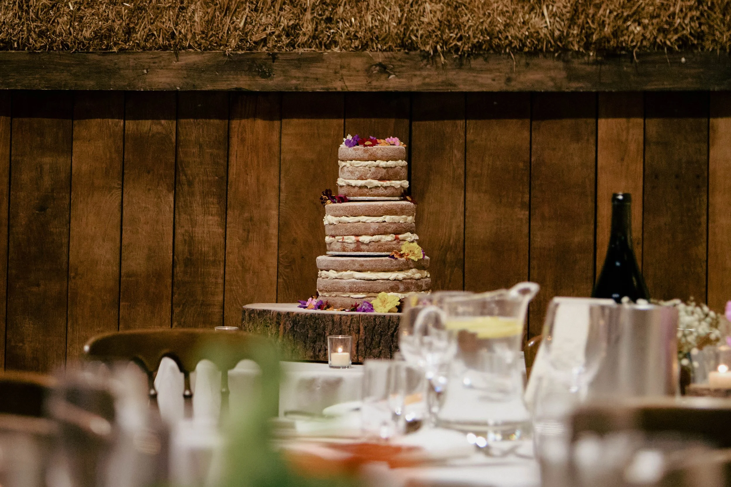 A tower of layered naked cakes with cream filling, decorated with edible flowers, placed on a tree trunk slab on a table at a rustic-style event.