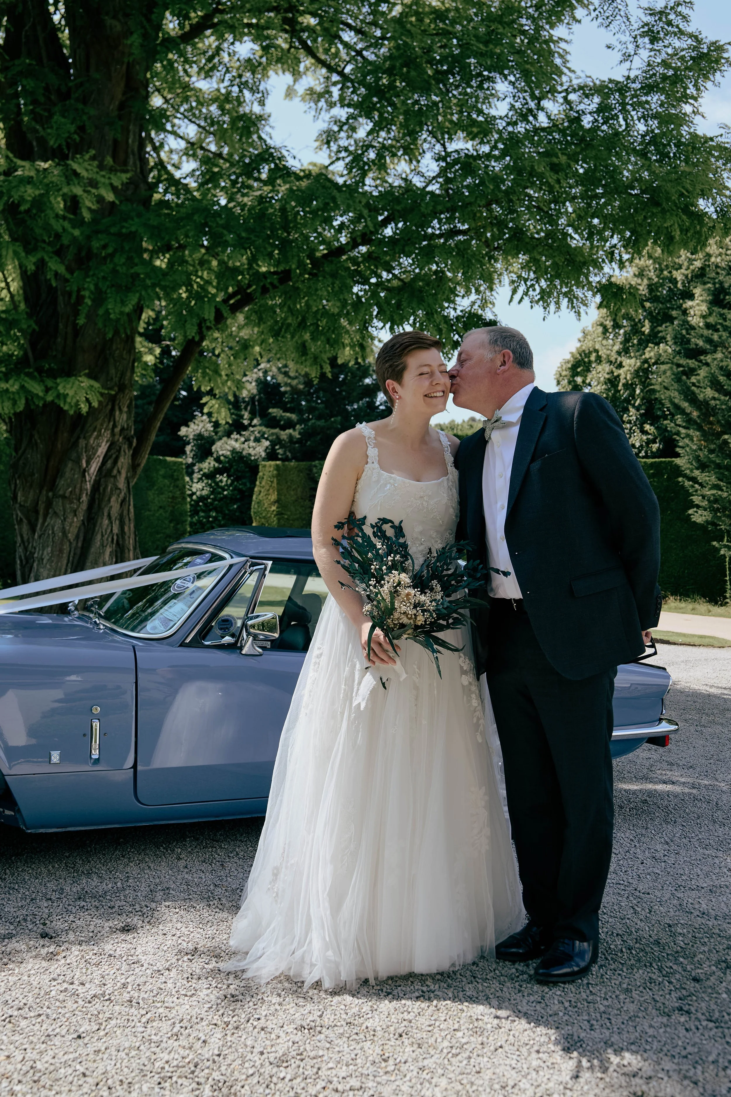 A bride and her father share a kiss and smile at each other outdoors during a wedding celebration. The bride wears a white wedding dress and holds a bouquet, the man is dressed in a dark suit with a bow tie. A classic blue convertible car is parked n