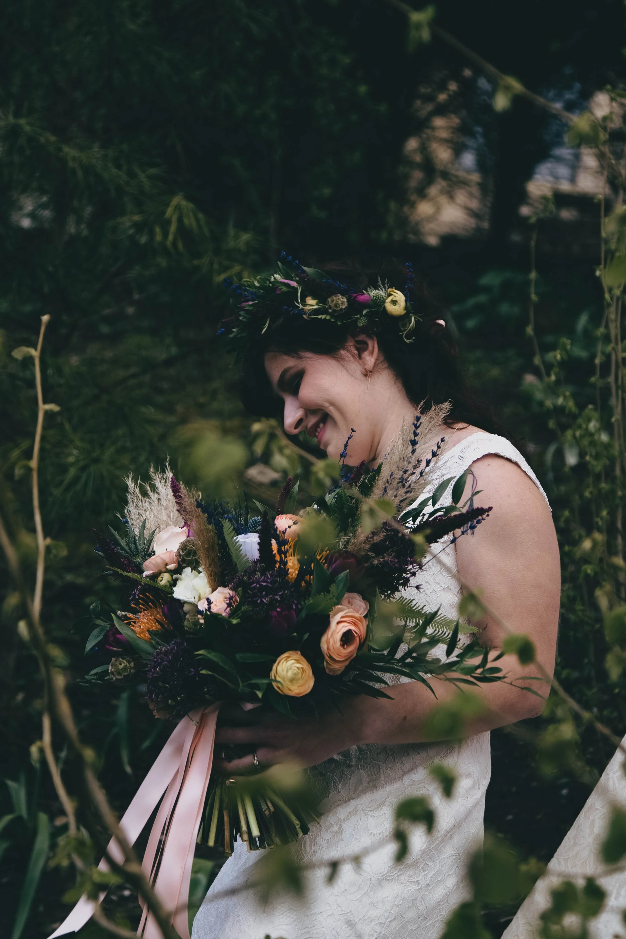 A woman in a white dress holding a large bouquet of flowers with greenery, roses, and other colorful blooms, standing outdoors among trees, wearing a flower crown, smiling and looking down.