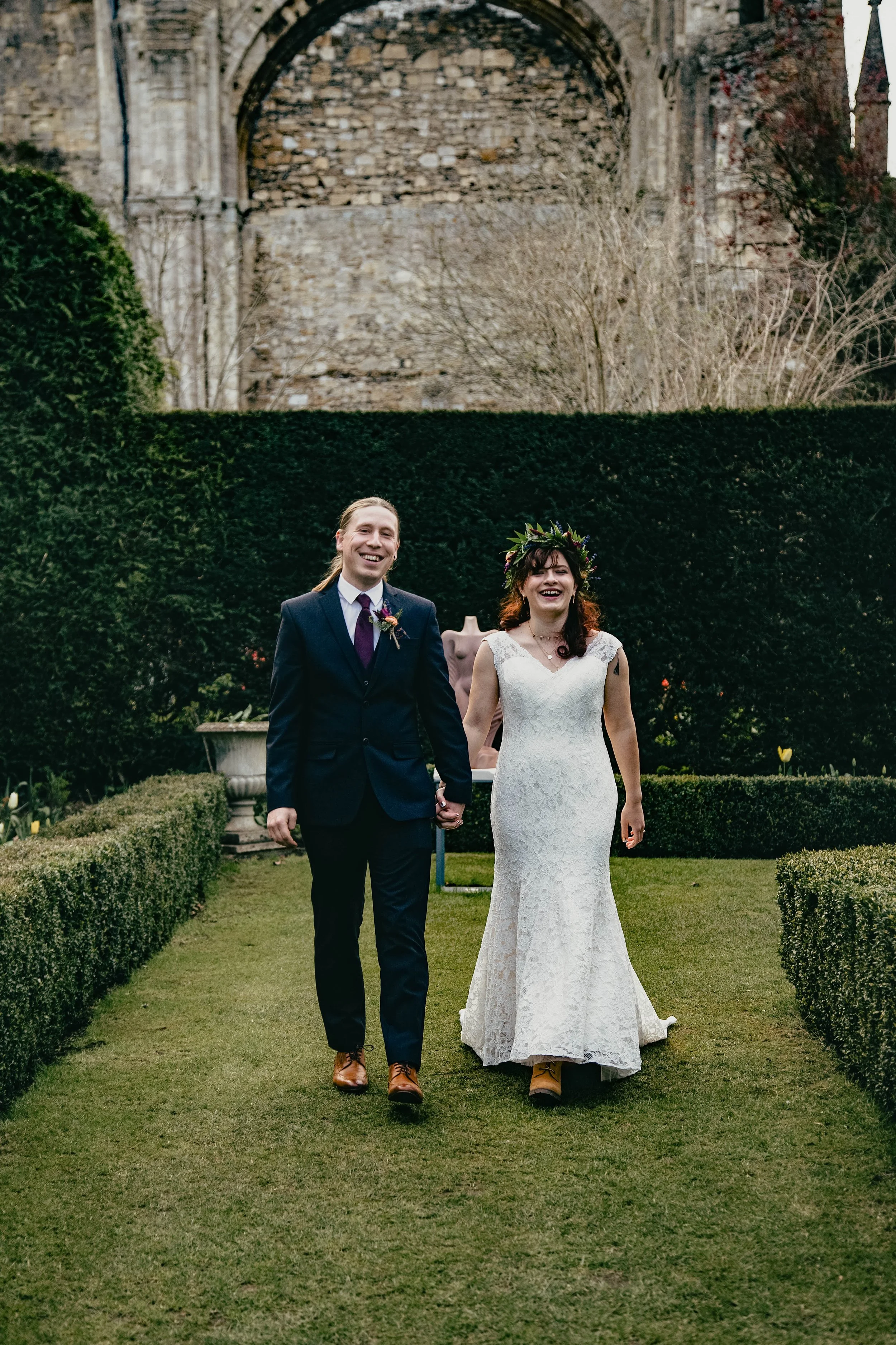 A couple in wedding attire holding hands and smiling in a garden with neatly trimmed grass and hedges, with Malmesbury Abbey in the background.