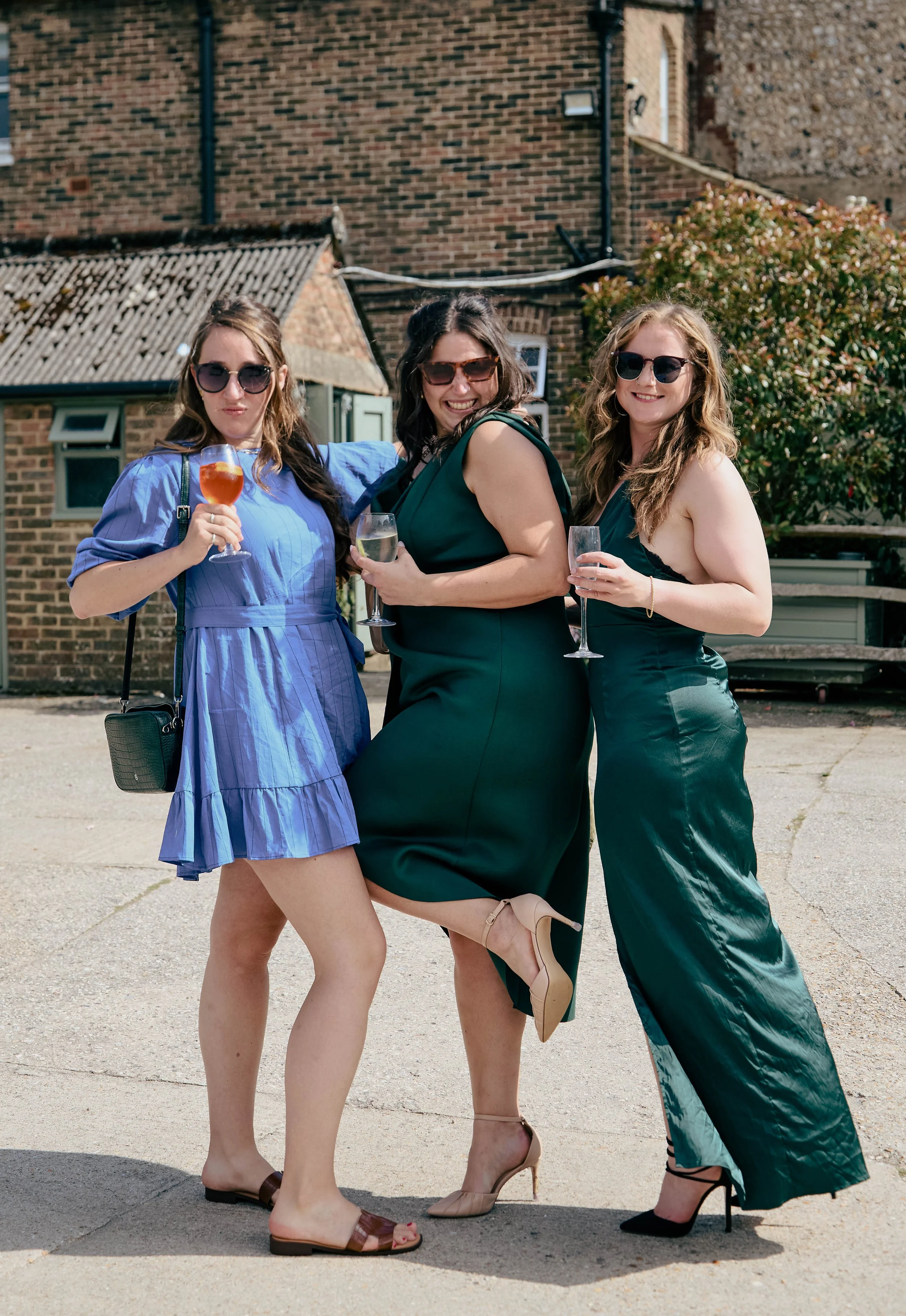 Three women dressed in elegant clothing, wearing sunglasses and holding drinks, standing outdoors against a brick building and bush, smiling and celebrating.