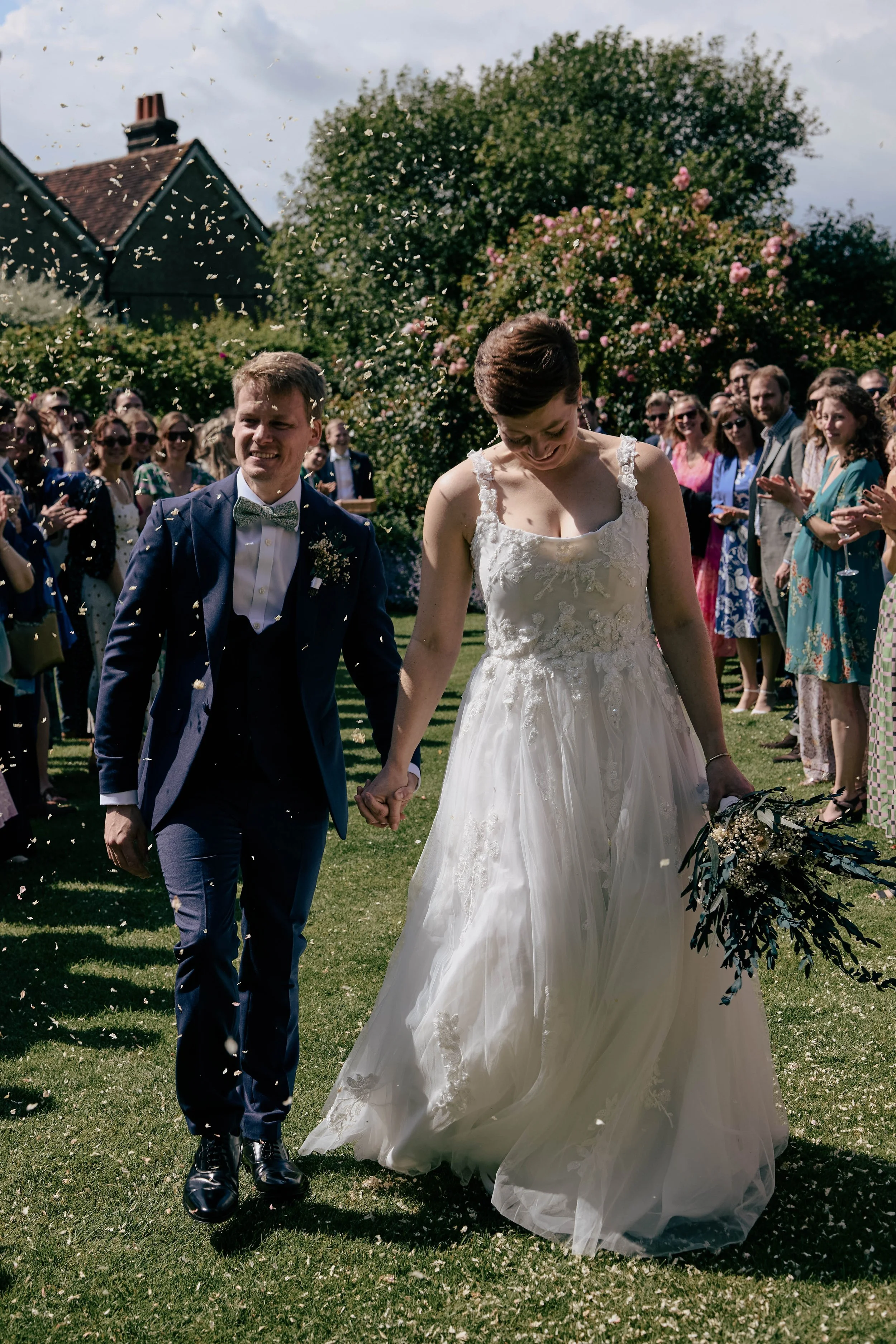 A bride and groom holding hands walking on a grassy lawn after their wedding ceremony, surrounded by friends and family clapping and celebrating, with confetti in the air and a house and blooming trees in the background.