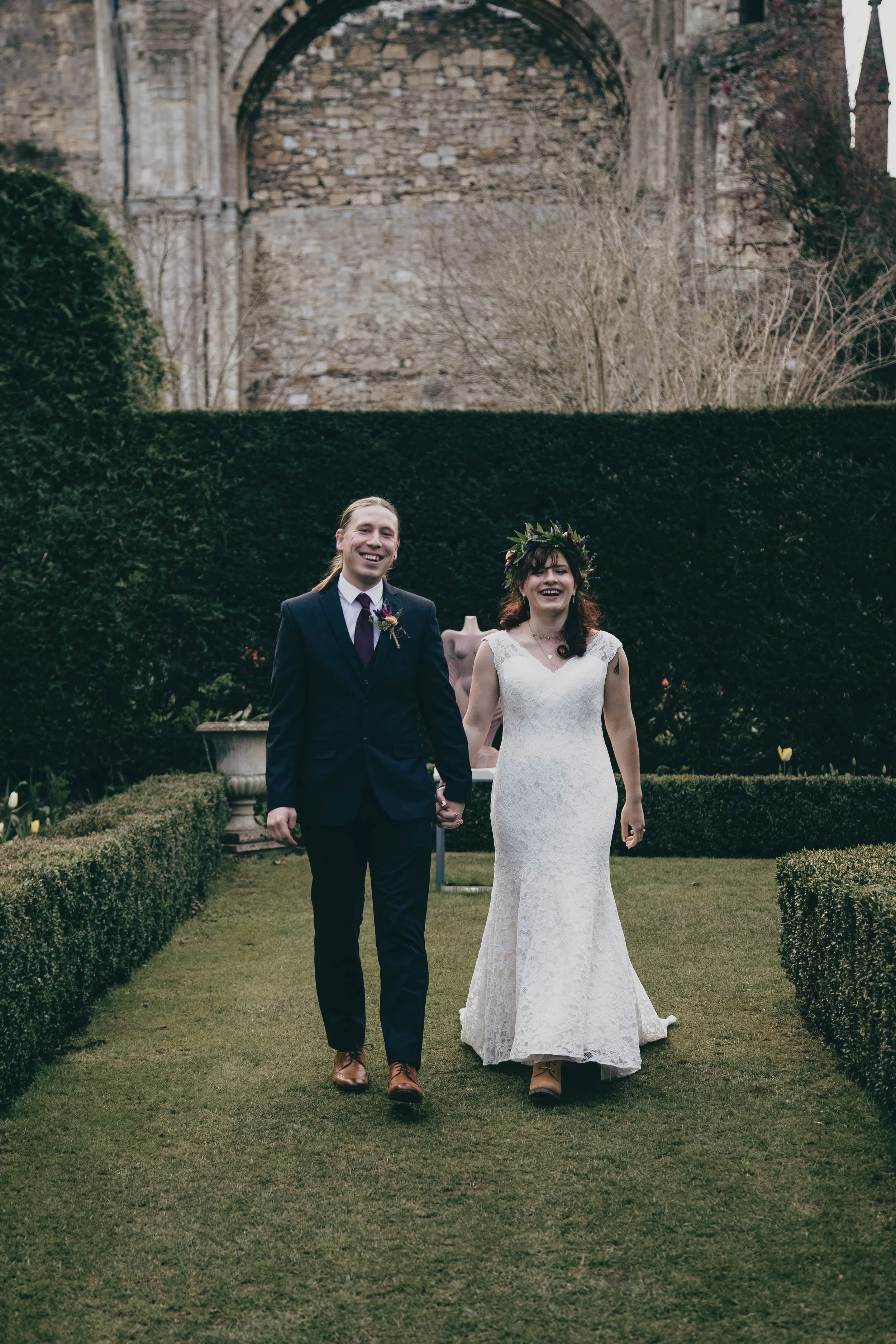 A newly married couple holding hands and smiling walking outdoors on a groomed lawn with a large stone wall and trees in the background at Malmesbury Abbey, Wiltshire.