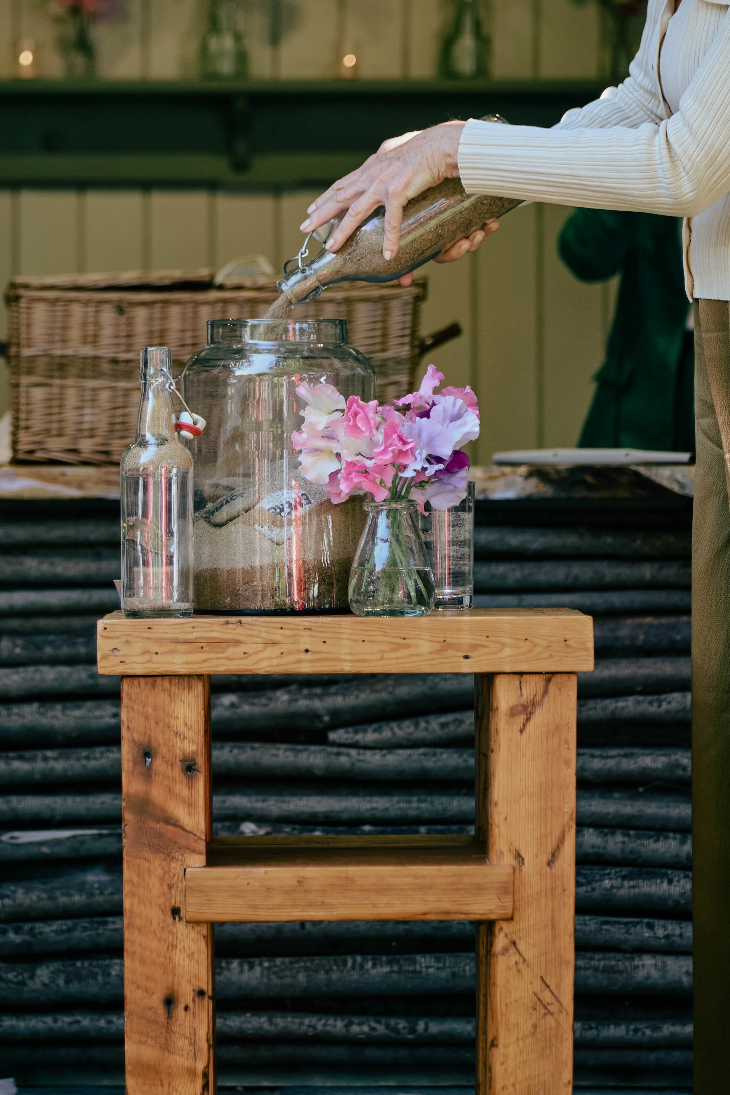 Someone pouring sand from a glass bottle into a large glass jar on top of a wooden table, with a smaller glass bottle and a vase of pink flowers nearby.