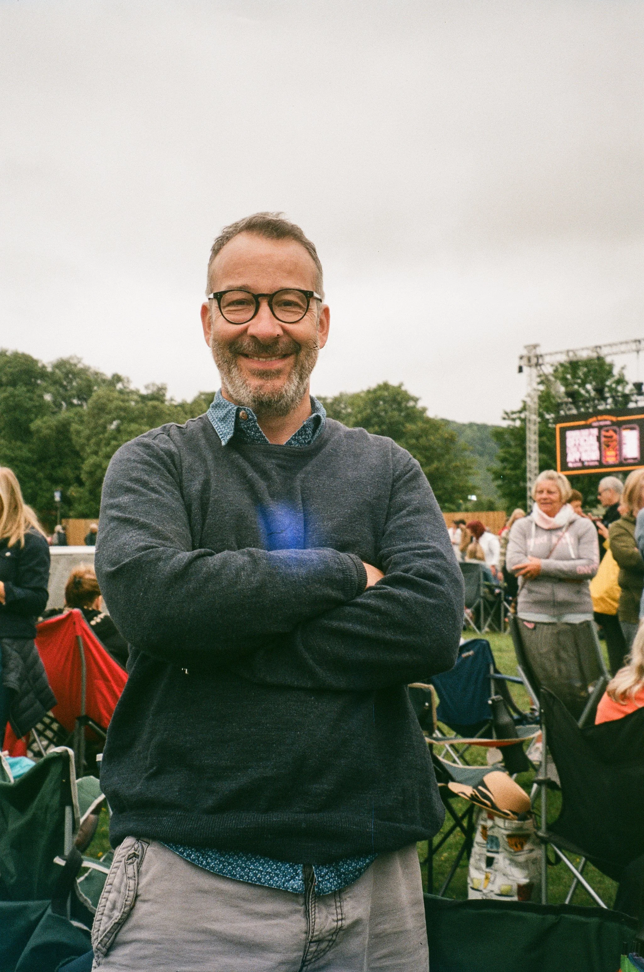 A smiling man with glasses and a beard wearing a dark sweater, standing outdoors at a gathering with other people, trees, and a digital scoreboard in the background.
