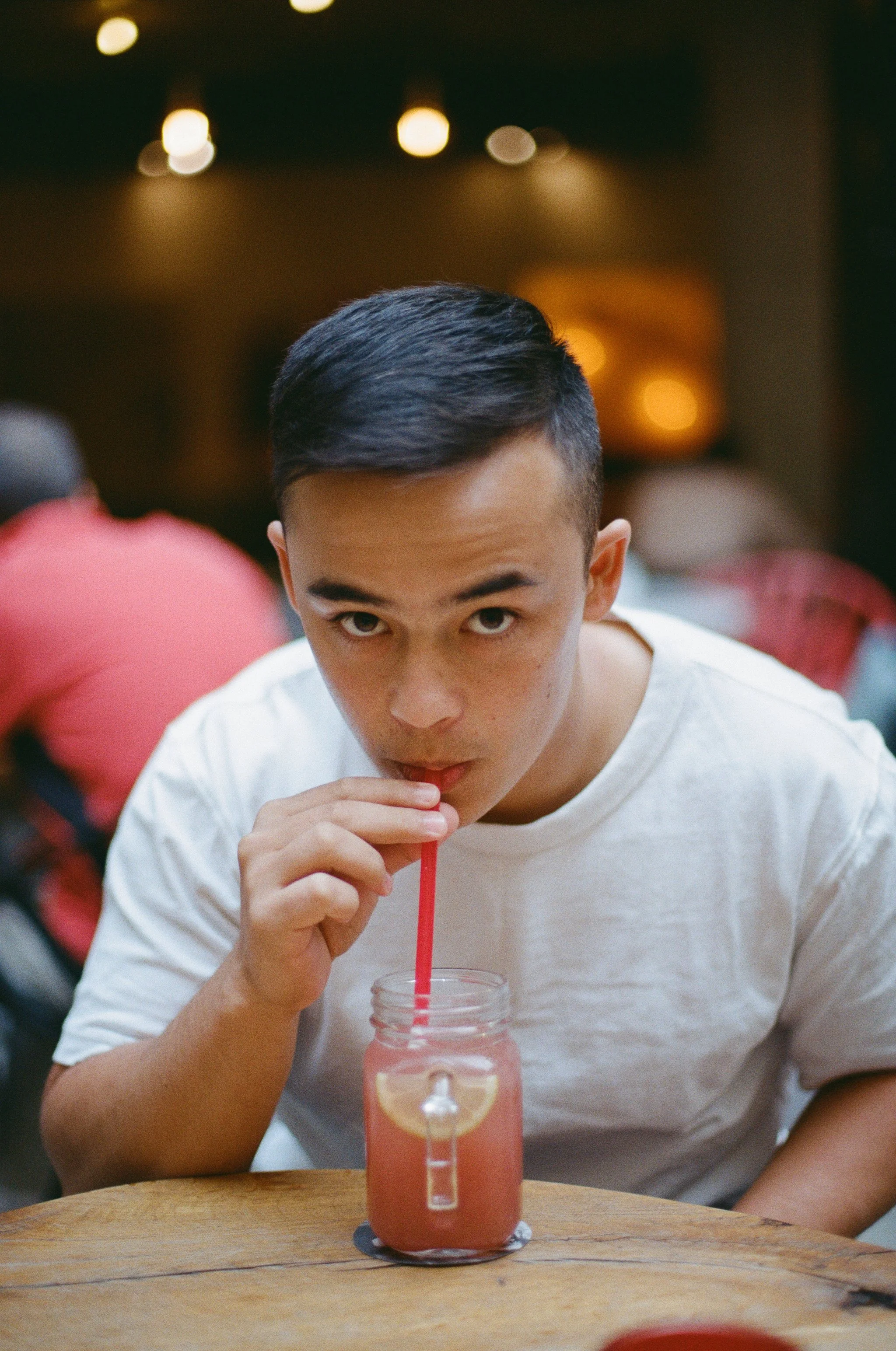 A young man with short, dark hair and a white shirt is sitting at a wooden table, drinking a pink beverage with lemon slices through a red straw. The background is dimly lit and slightly blurred, with abstract circular lights.