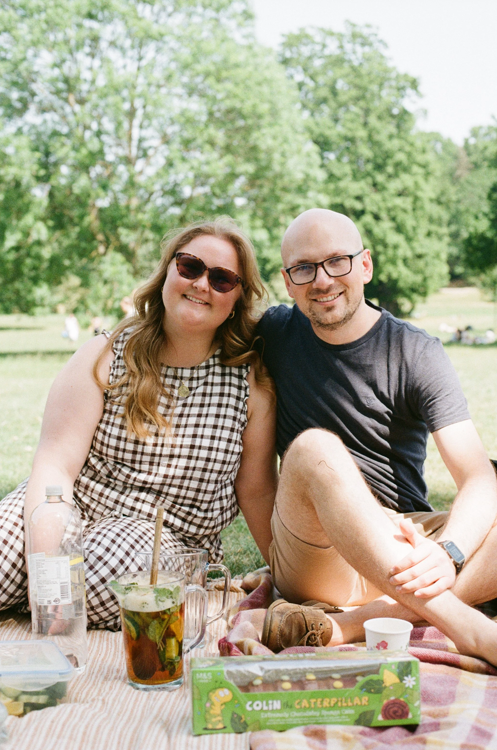 A couple sitting outdoors during a picnic, smiling at the camera, with food and drinks on a blanket in front of them, surrounded by green trees.