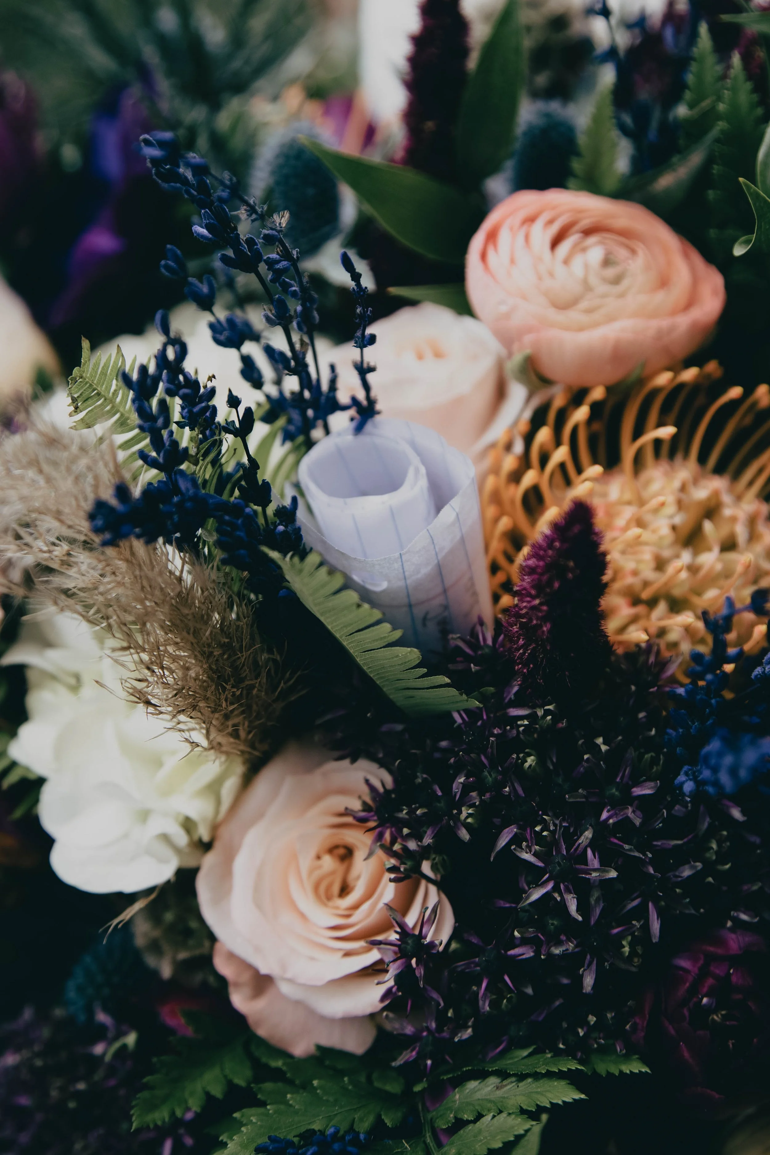 A bouquet of various flowers, including pink ranunculus, purple lavender, white roses, and green foliage, with a note hidden in the bouquet.