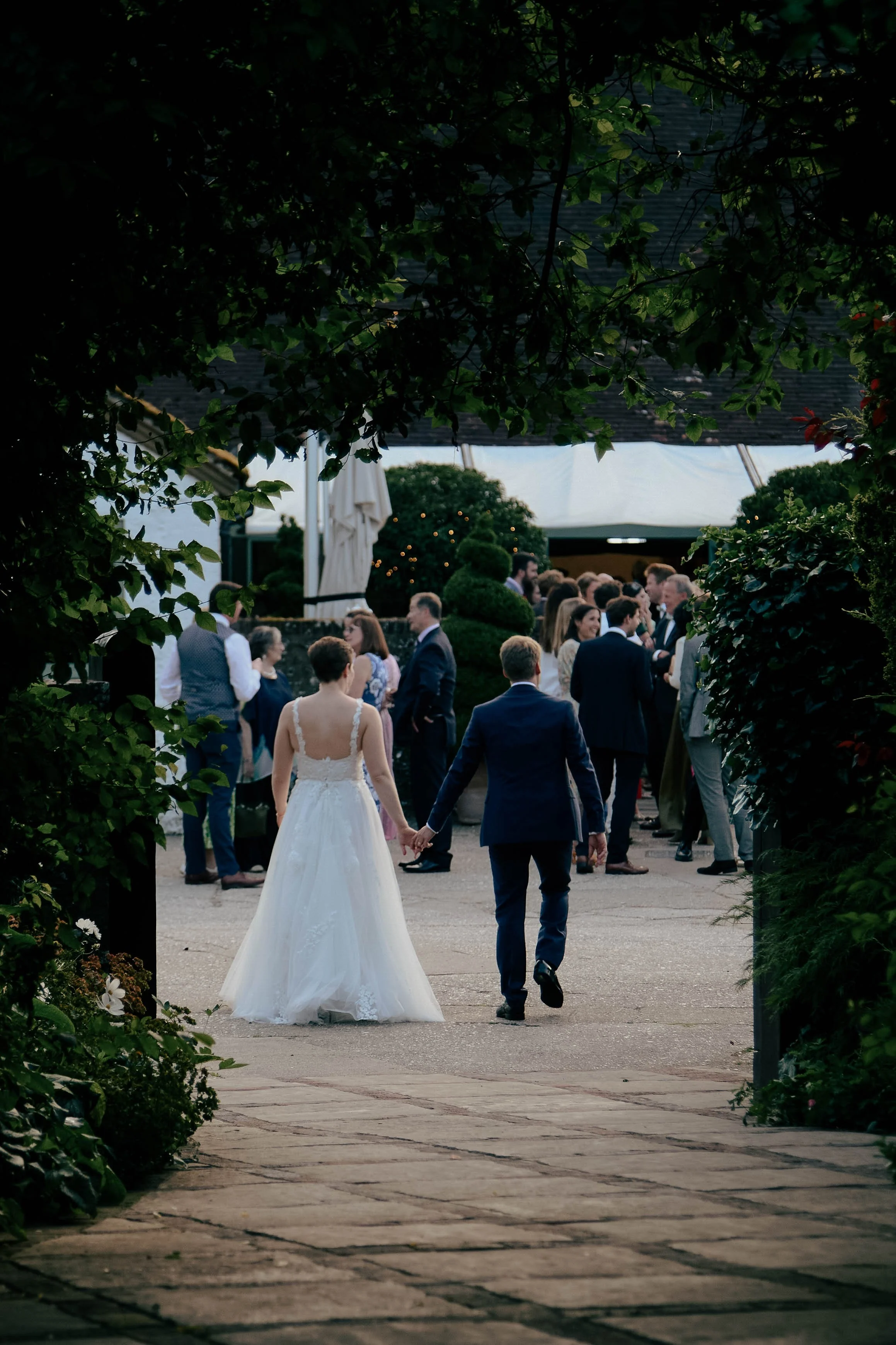 A bride and groom walking hand in hand at an outdoor wedding reception, seen through a framed opening of lush green leaves.