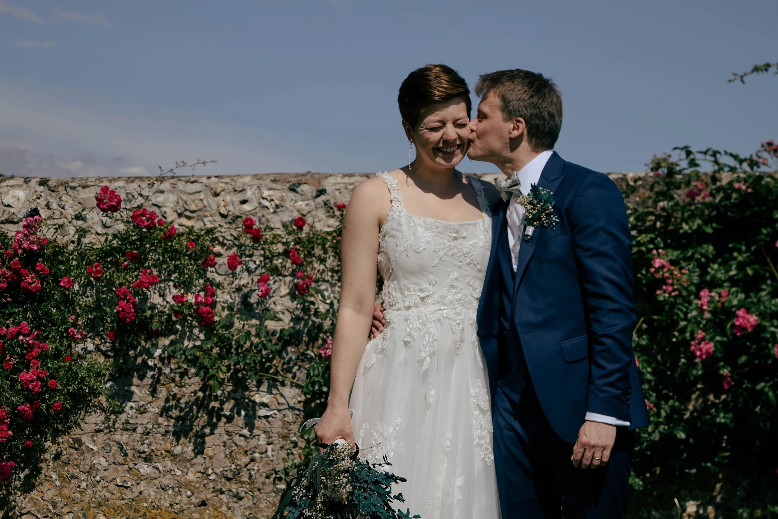 A bride and groom sharing a kiss, with the groom giving the bride a kiss on the cheek, outside in front of a stone wall with pink flowers and a blue sky.