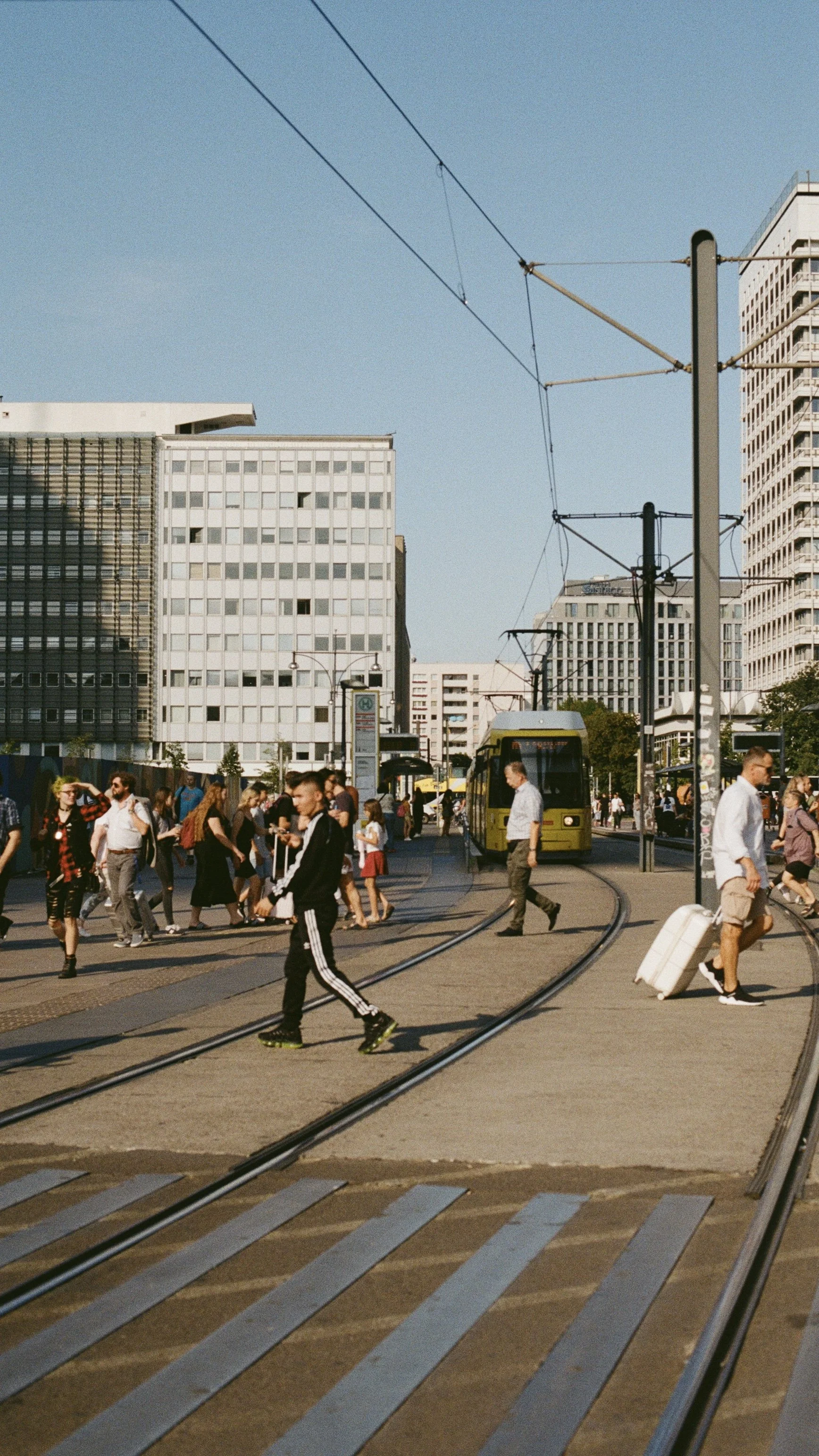 A busy city street with pedestrians crossing at a tram stop in Berlin, a yellow tram on tracks, and tall office buildings under a clear blue sky.