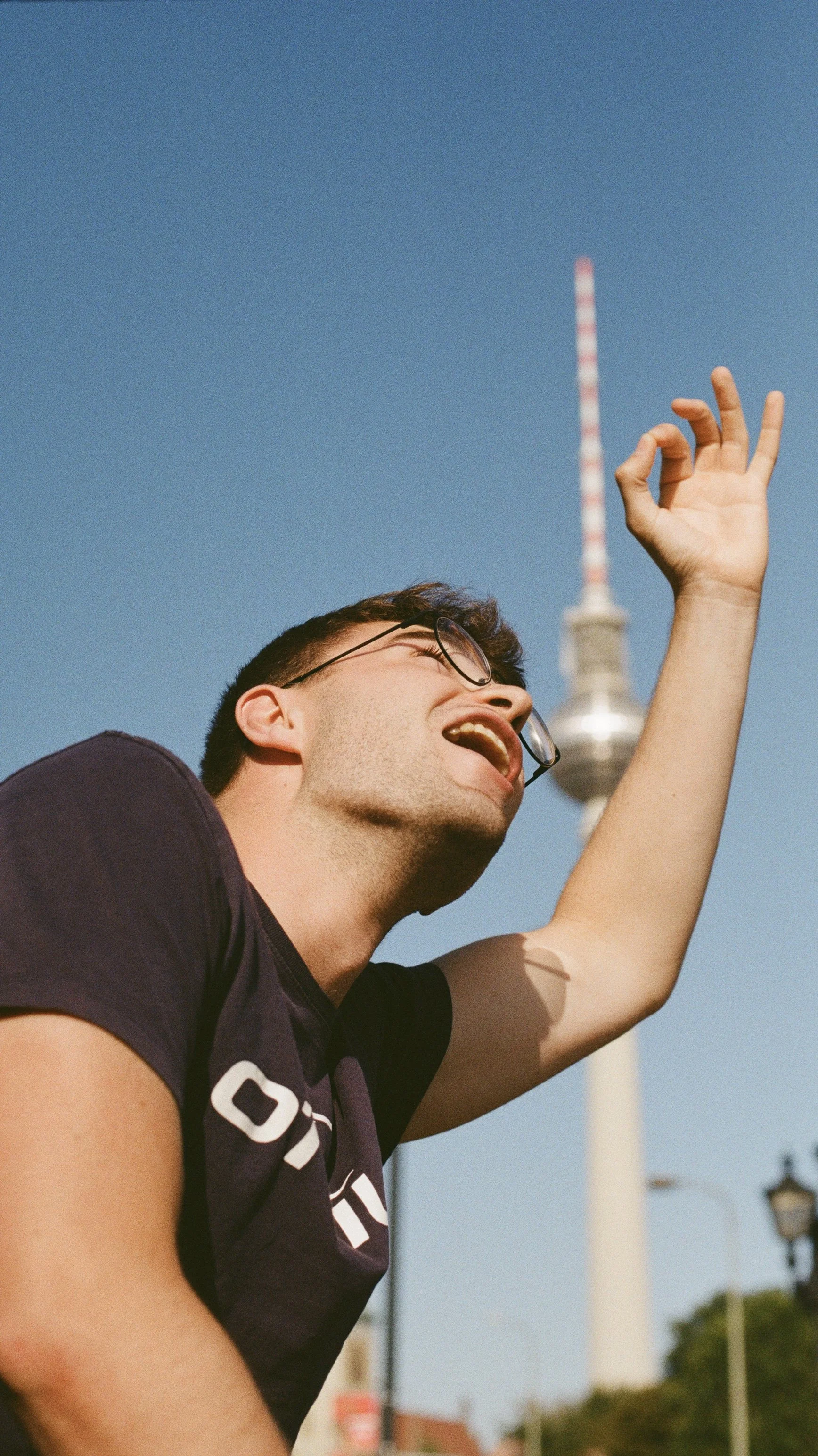 A young man with glasses and a dark T-shirt smiling and making an OK gesture with his hand, against a clear blue sky with the Berlin TV Tower in the background.