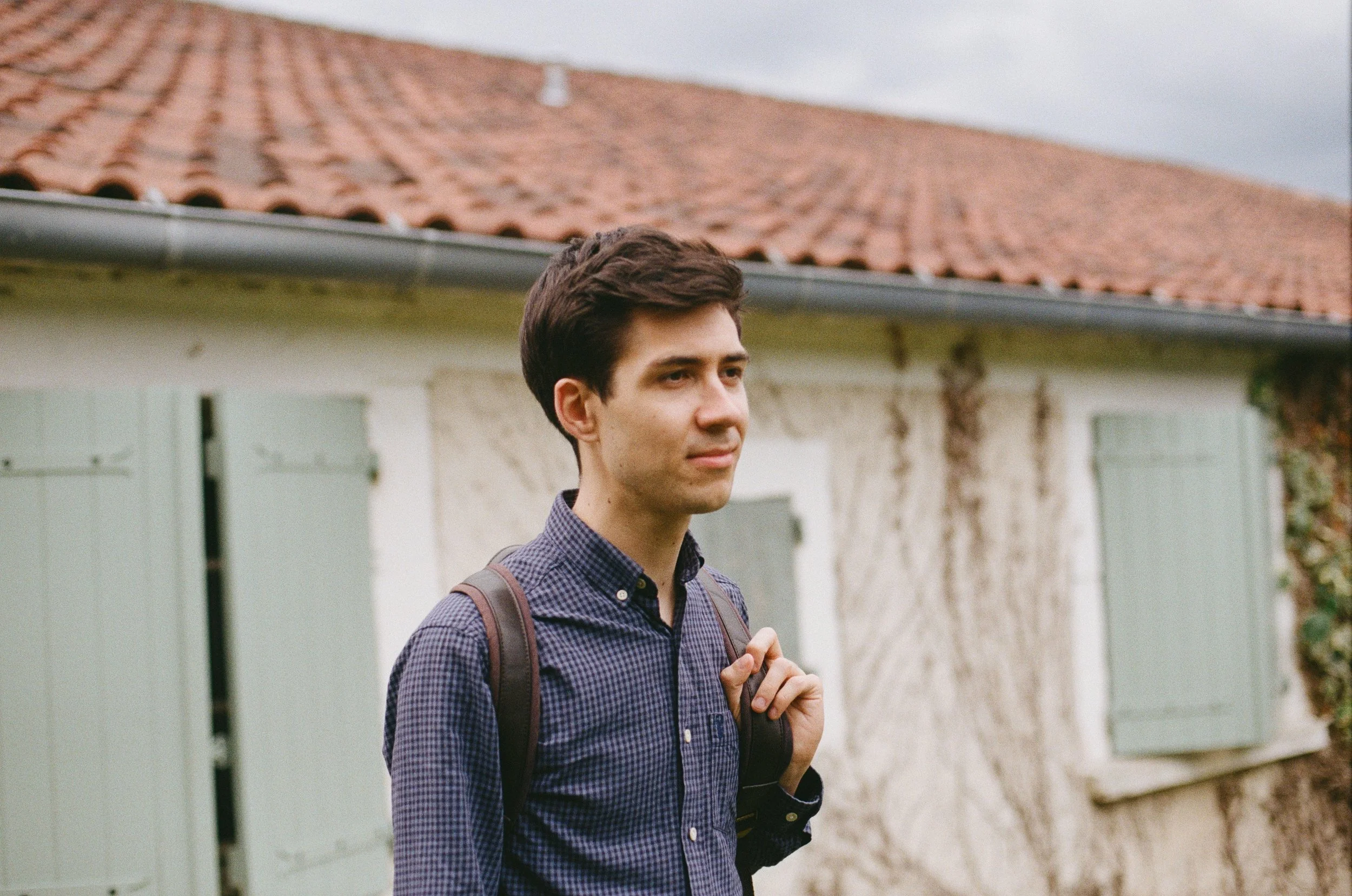 A young man with dark hair, wearing a checkered shirt and carrying a backpack, standing outdoors near a building with closed shutters and a tiled roof.