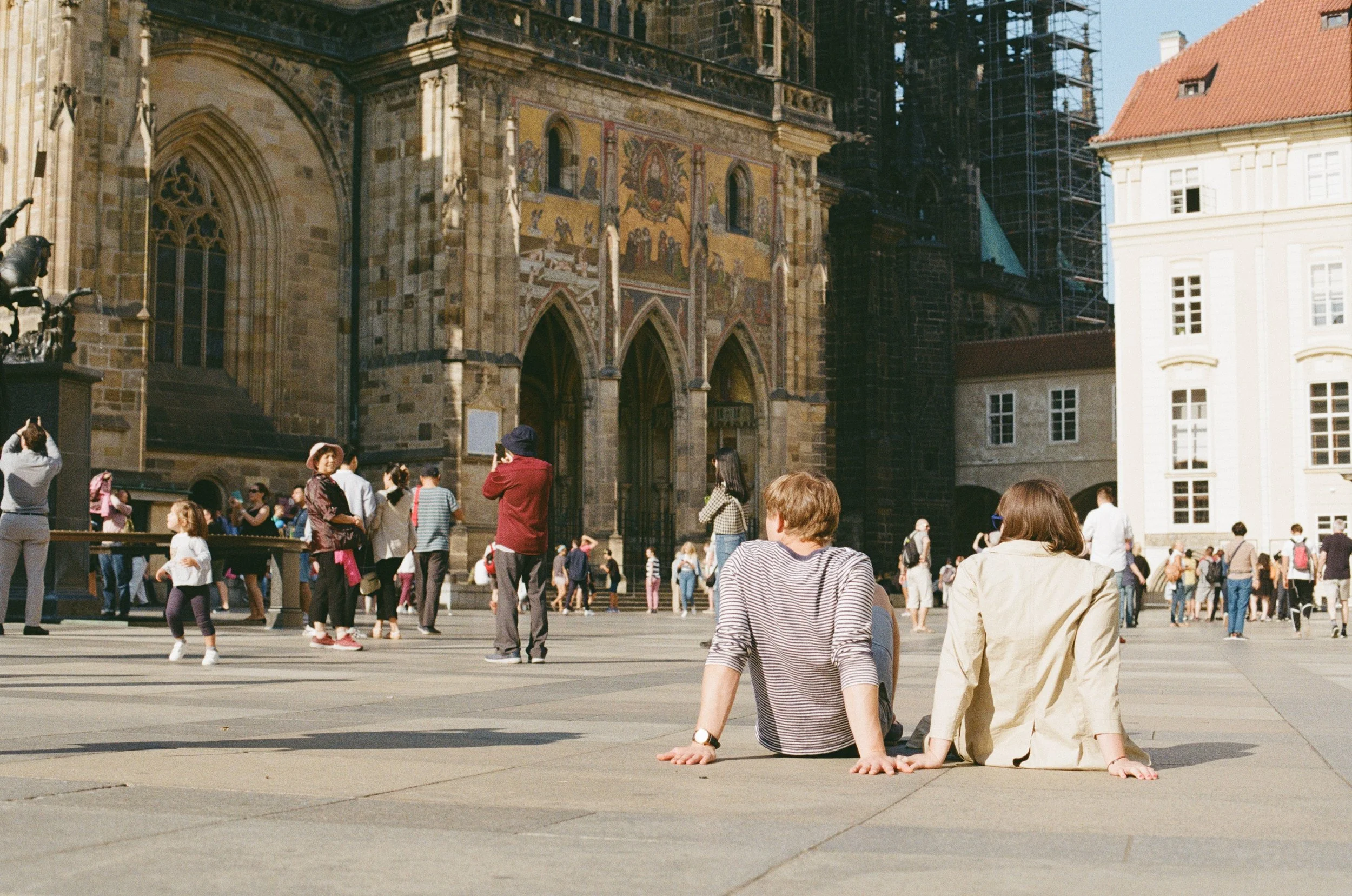 People gathered in a square in front of a historic building with detailed stonework and murals, a couple sitting on the ground subtly touching hands.