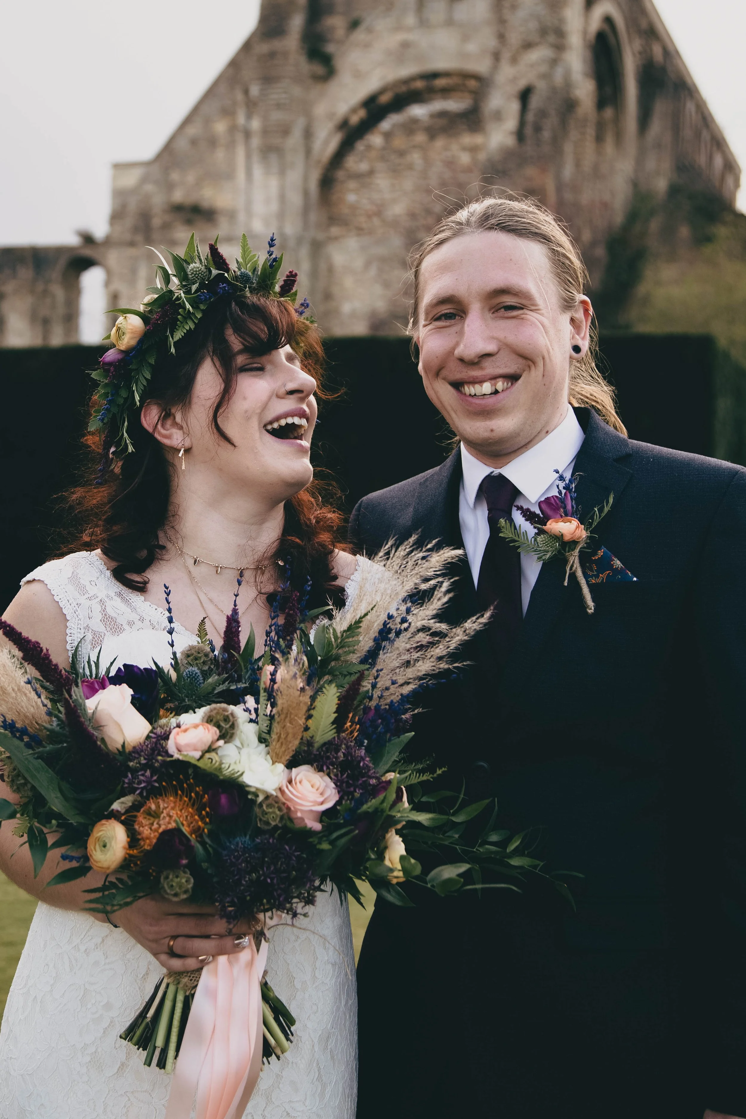 A bride and groom smiling at a wedding. The bride is holding a large bouquet of colorful flowers, and the groom is dressed in a dark suit with a boutonniere. They are outdoors with what appears to be an ancient stone building or ruins in the backgrou