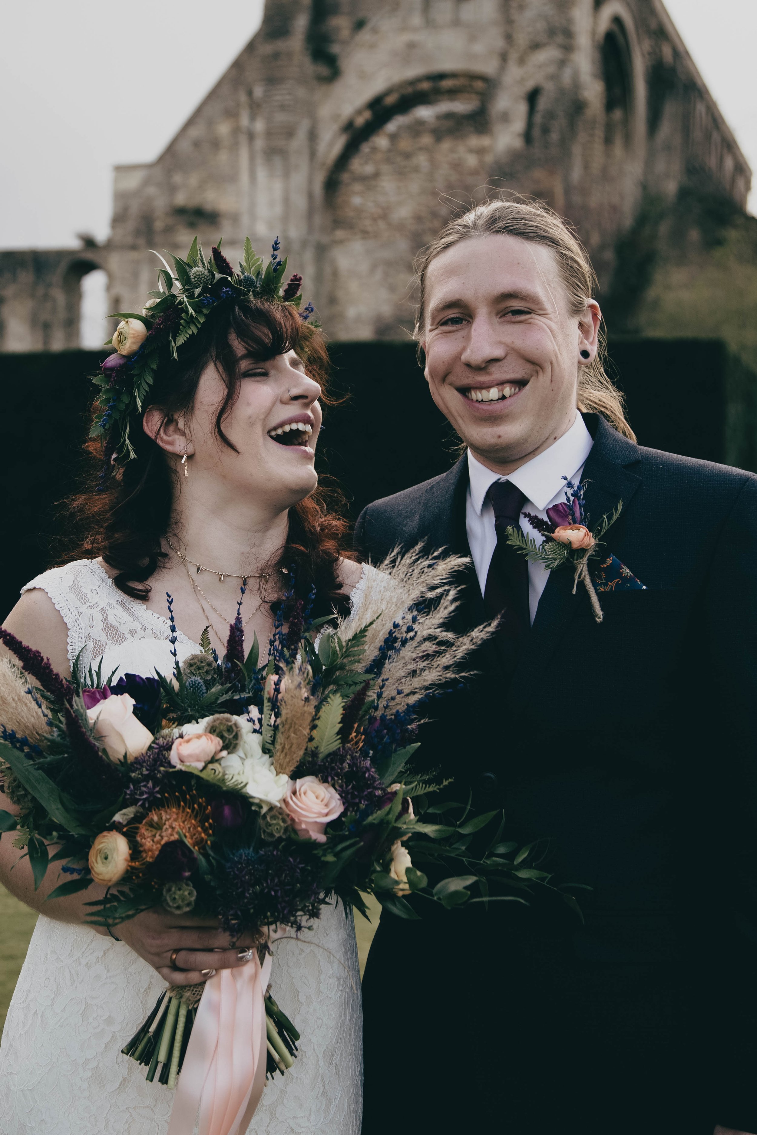 A smiling couple dressed in wedding attire, with a woman holding a large bouquet of flowers, standing outdoors near an old stone building (Malmesbury Abbey, Wiltshire).