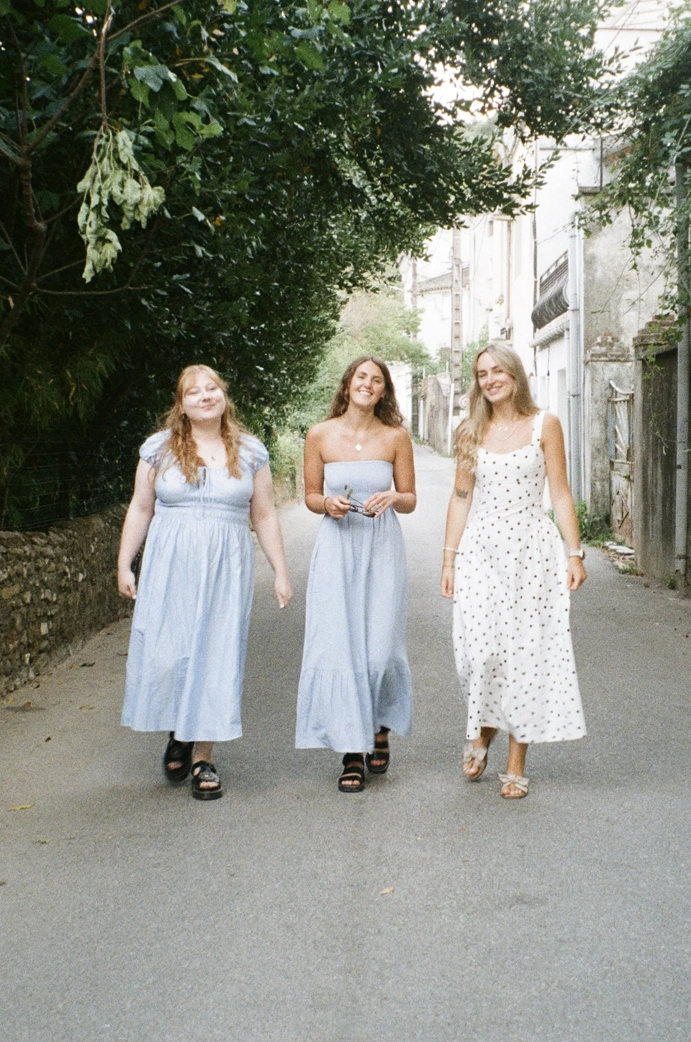 Three young women walking down a street lined with trees and buildings, smiling and wearing summer dresses.