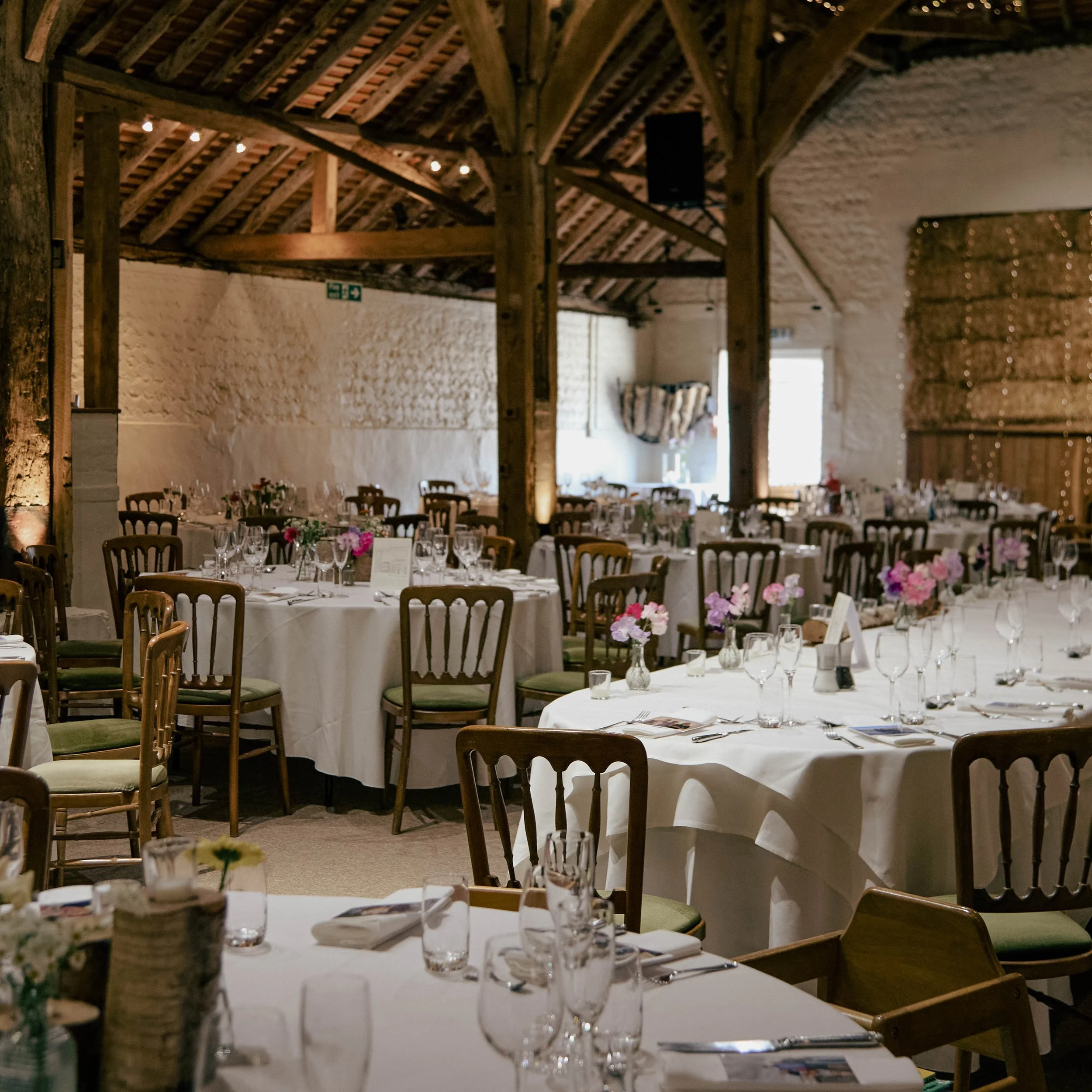 Interior of a rustic banquet hall with high wooden ceilings at Pangdean Old Barn, white brick walls, and round tables set with white tablecloths, glassware, and pink flowers in small vases, prepared for an event. 