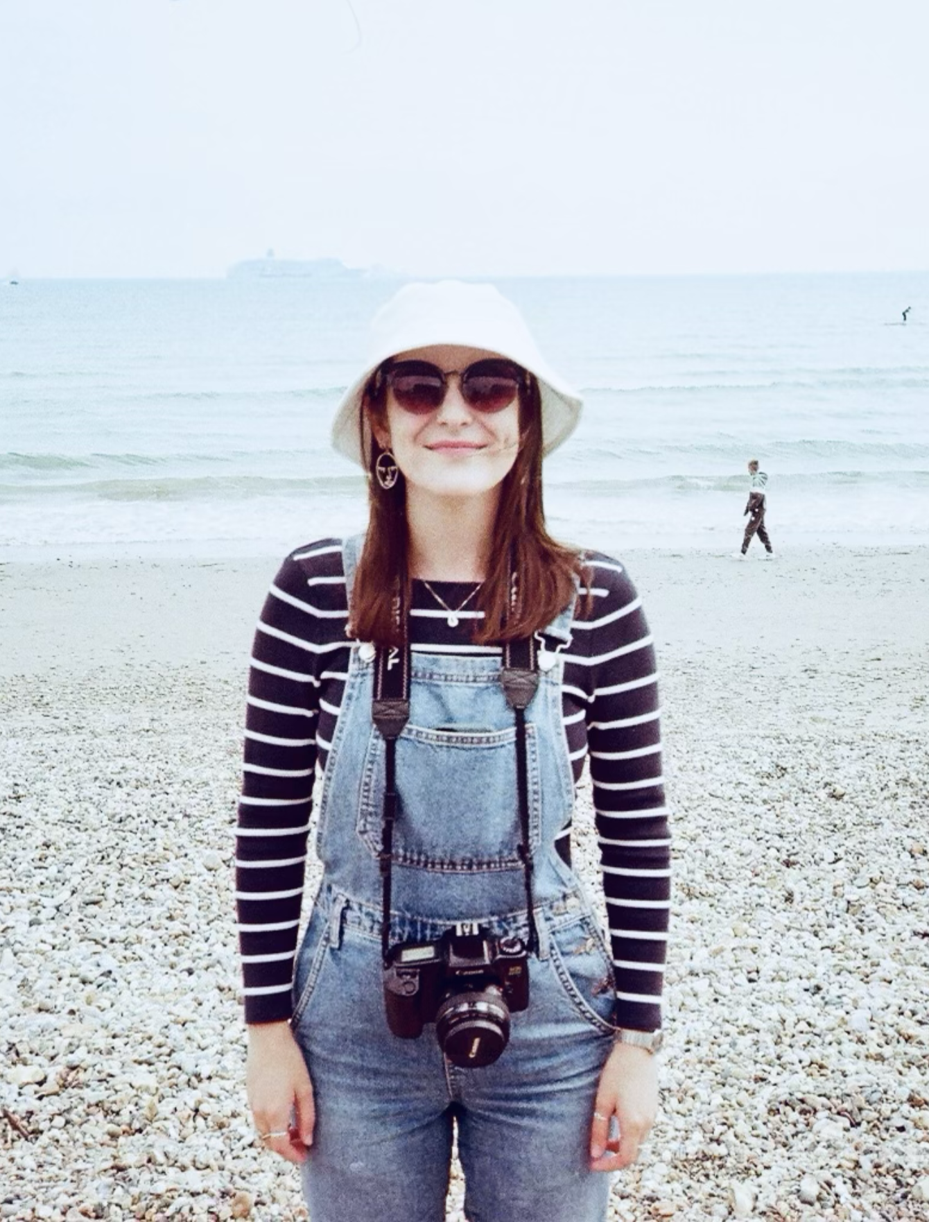 A woman wearing sunglasses, a white hat, a striped shirt, and denim overalls standing on a pebble beach with the ocean behind her. In the background, a person is walking along the shore and a ship is visible on the water.