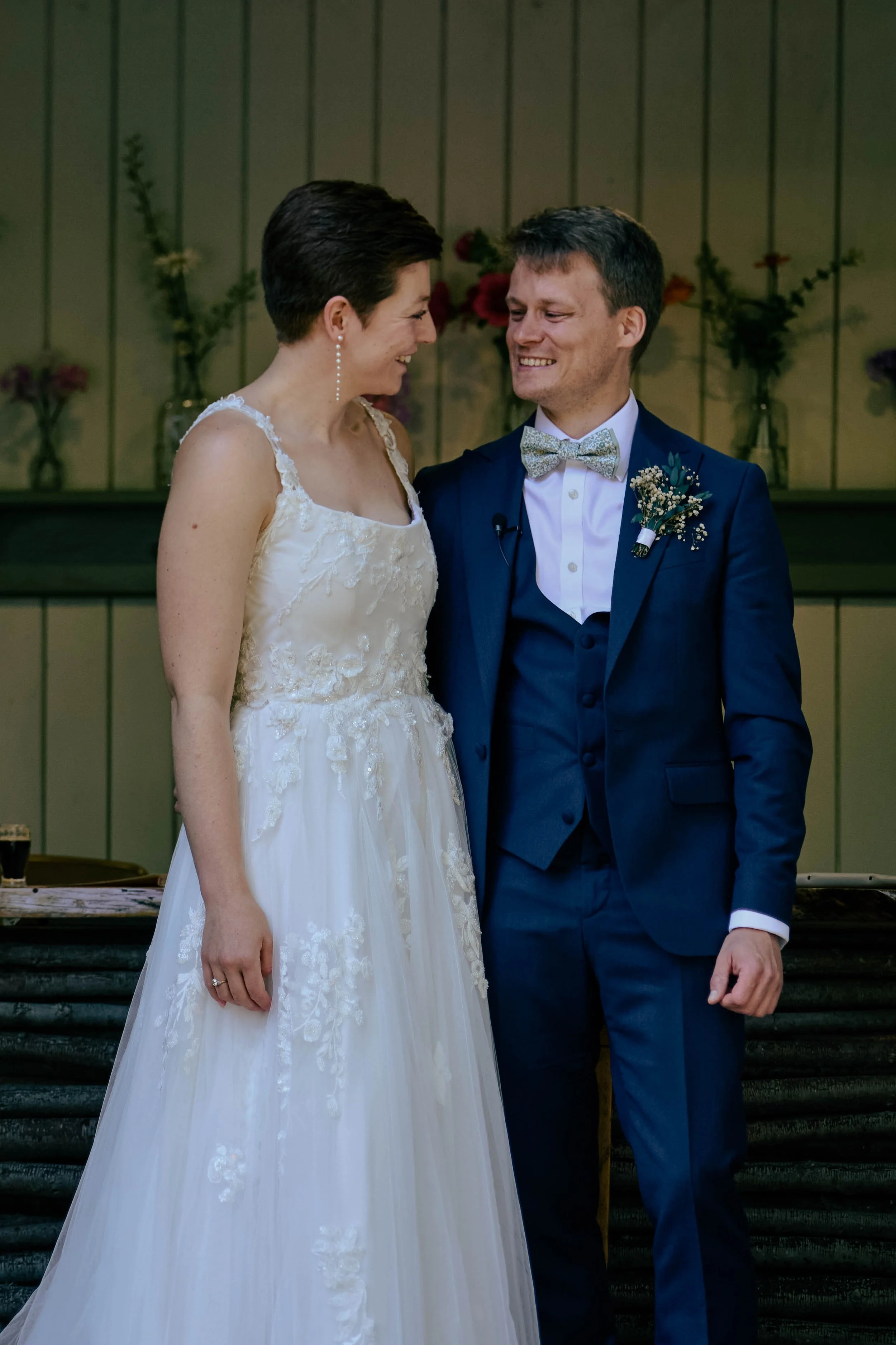 A bride and groom smiling at each other after their wedding ceremony, standing on stairs indoors with flower arrangements in vases in the background.