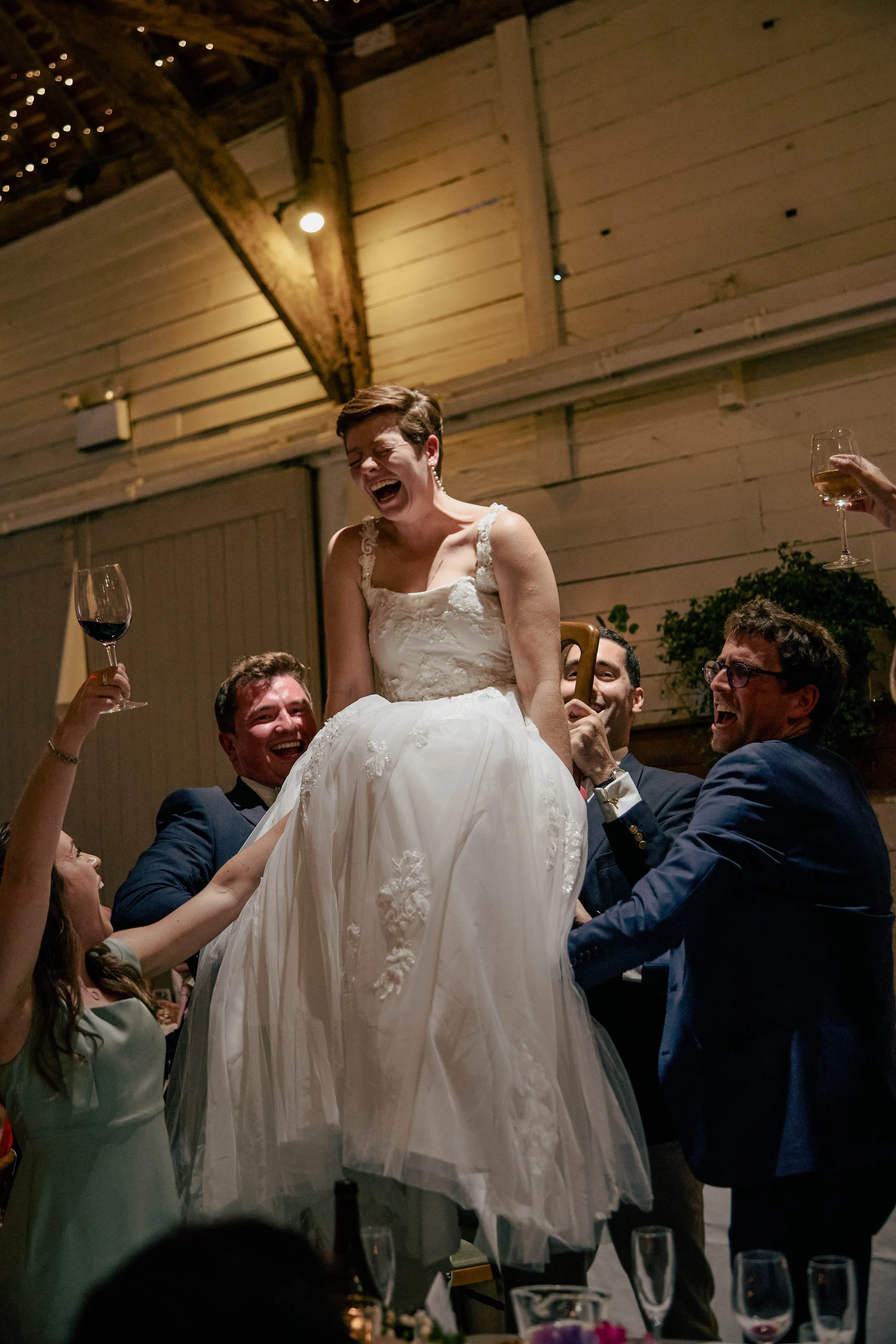 A woman in a wedding dress being lifted in the air by a group of people at a wedding reception. She is laughing, while others around her are smiling and also holding glasses.