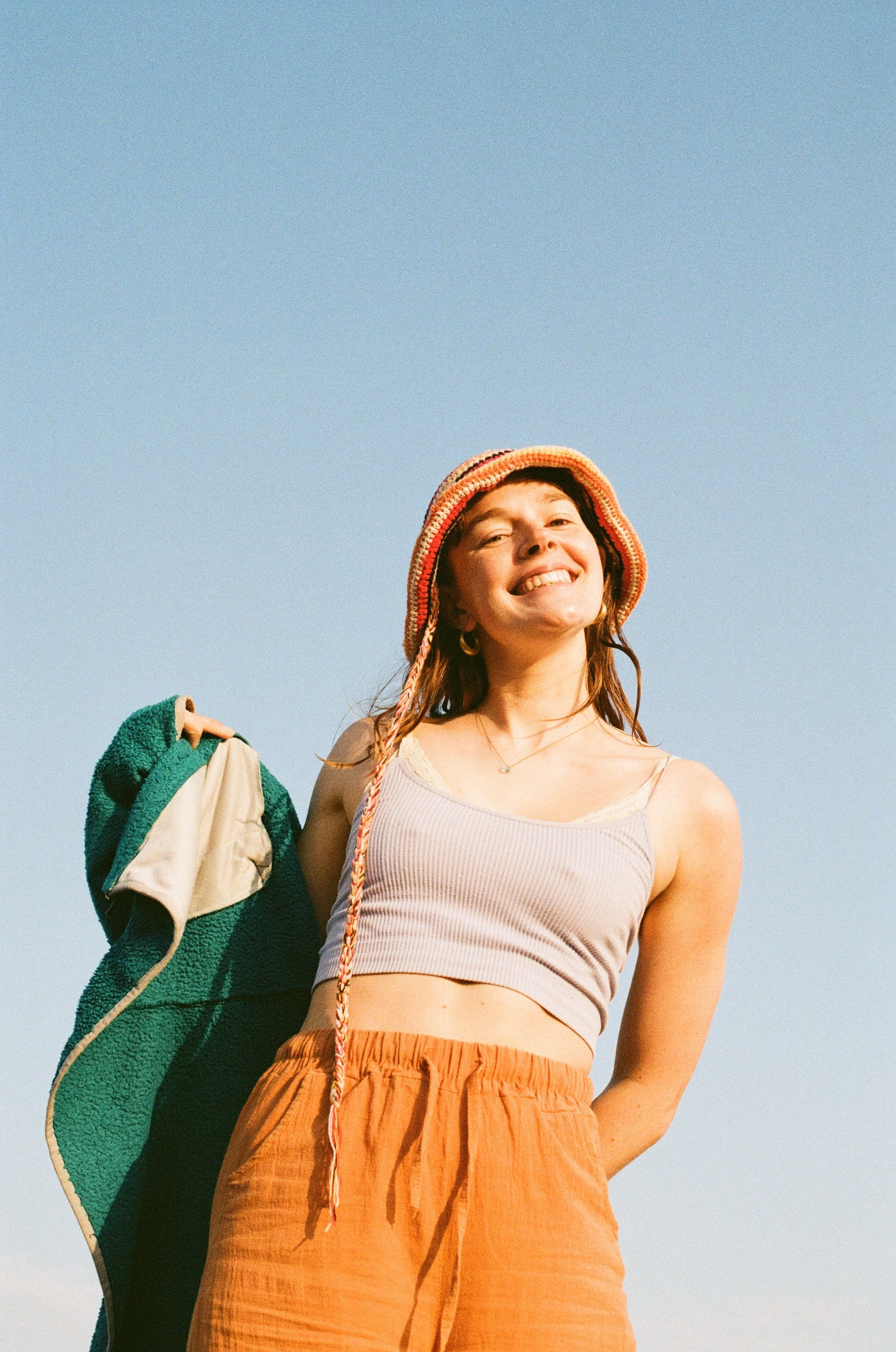 A young woman with a cheerful expression, wearing a knitted hat with colorful stripes, a beige tank top, and orange high-waisted pants, holding a green and beige jacket, standing outdoors against a clear blue sky.