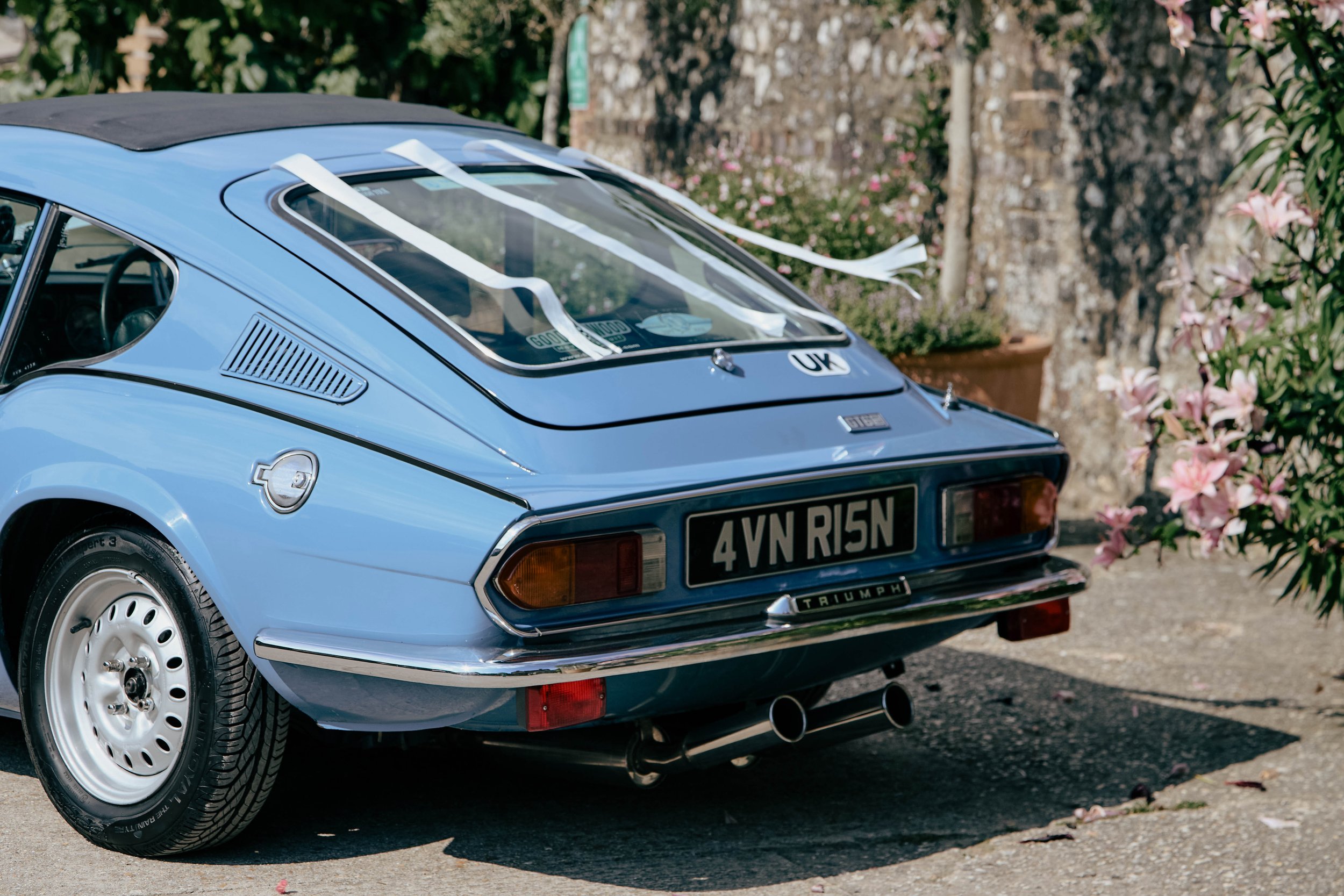 A blue classic Triumph sports car parked outdoors with pink flowers and green foliage in the background.