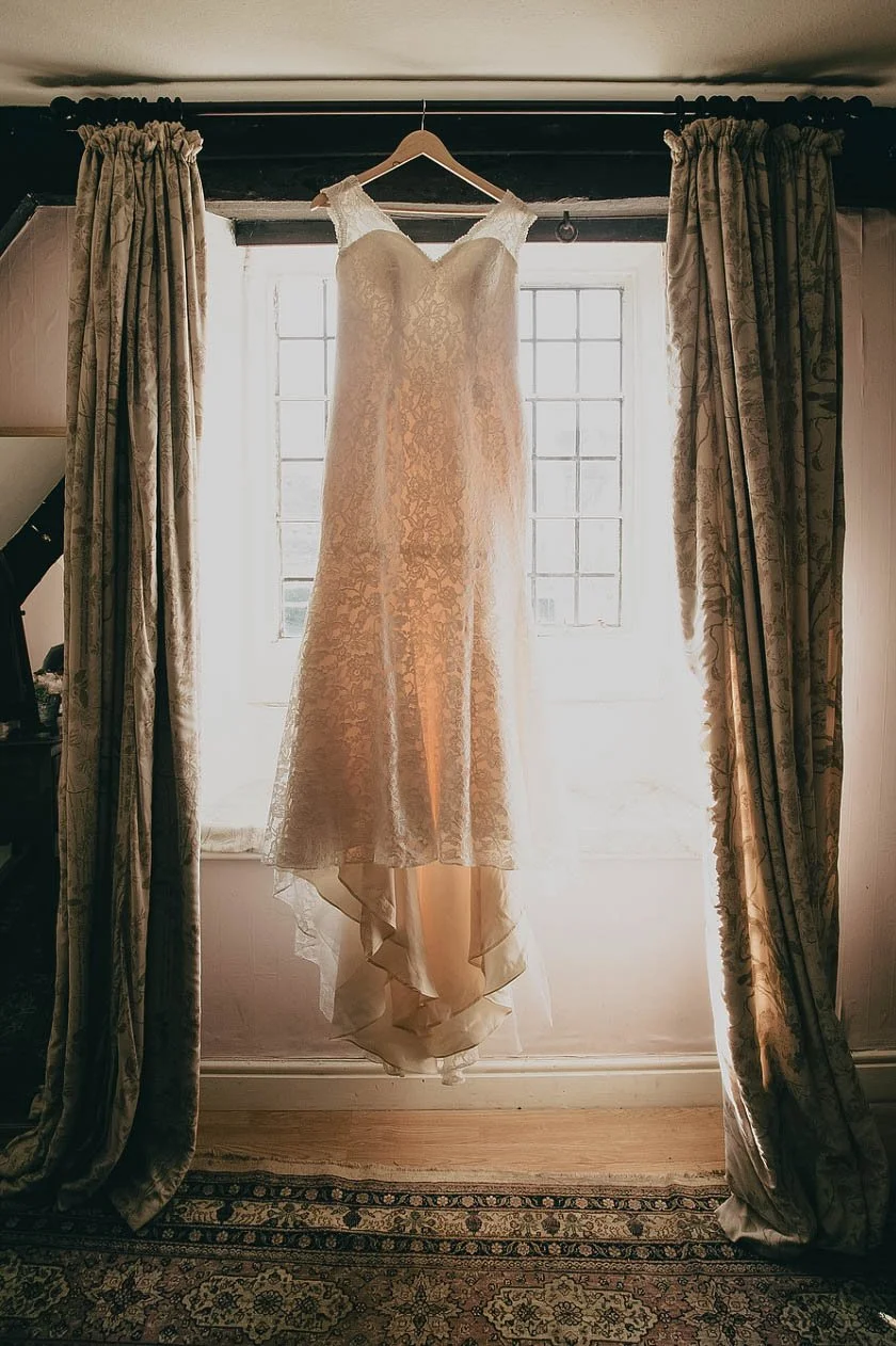 A wedding dress is hanging on a hanger in front of a window, with curtains on either side. The dress is made of lace and appears to be cream-colored.