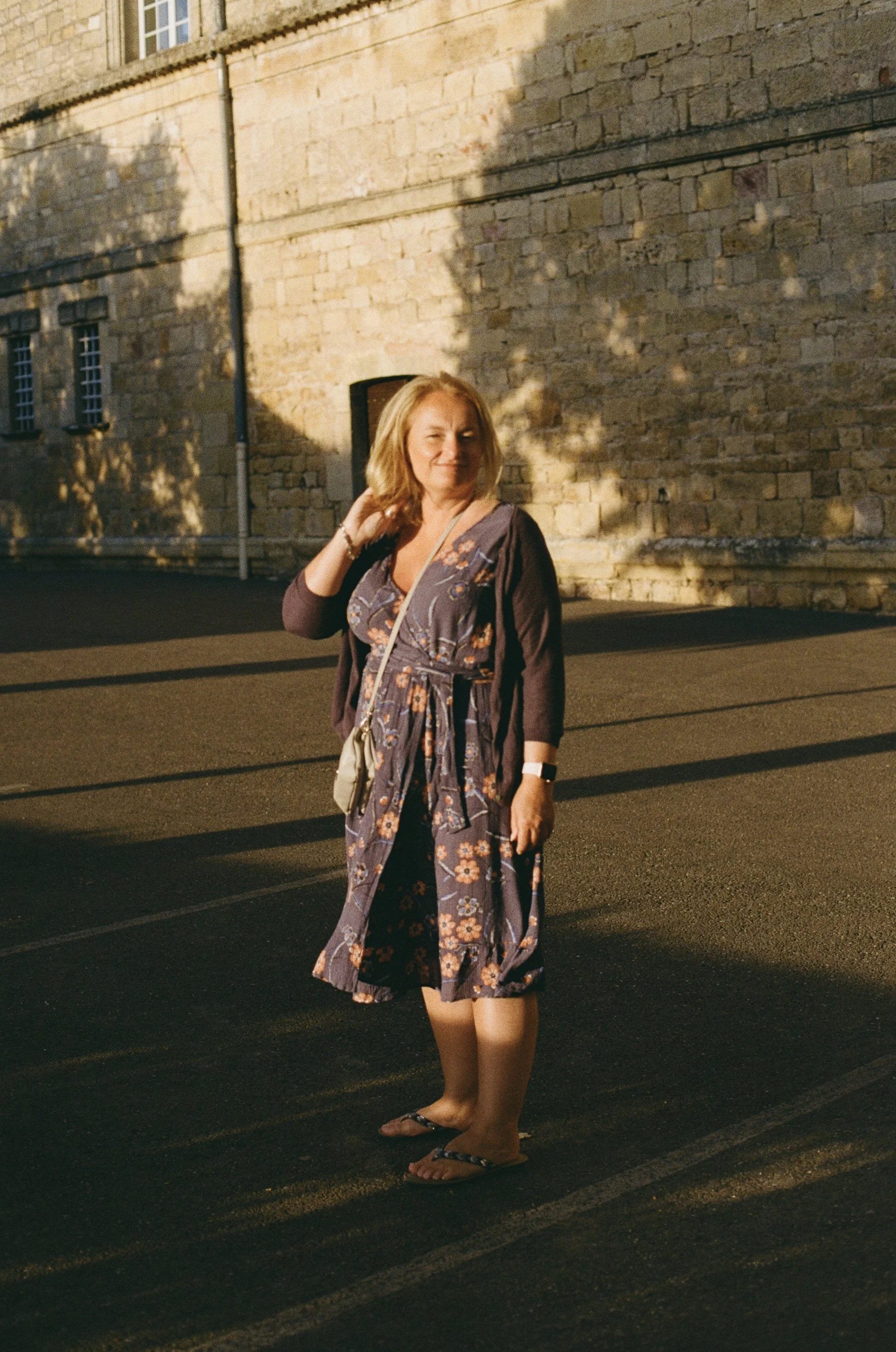 A woman with blonde hair wearing a floral dress and a dark cardigan, standing outside on the pavement near a stone building, in warm sunlight with shadows cast on the wall behind her.