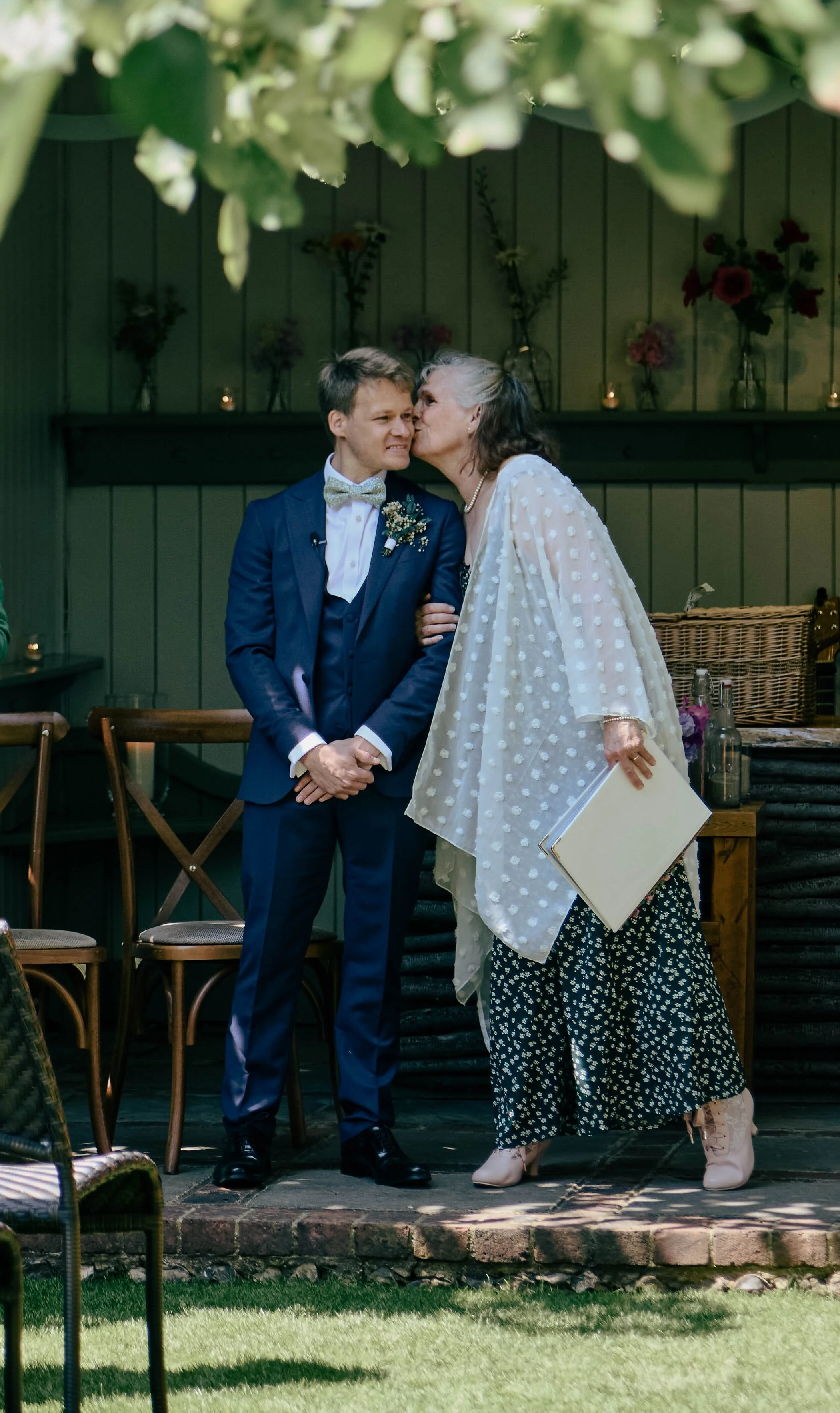 A woman giving a kiss on the cheek to a young man dressed in a suit, standing outdoors on a patio with grass in the foreground and flowers and candles in the background.