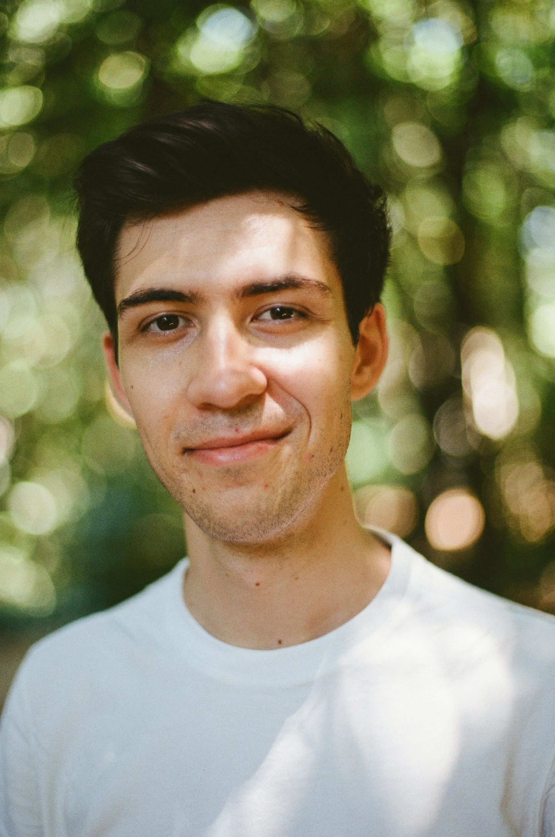 A young man with black hair and light skin, smiling slightly, wearing a white shirt, outdoors with blurred green and white bokeh background.
