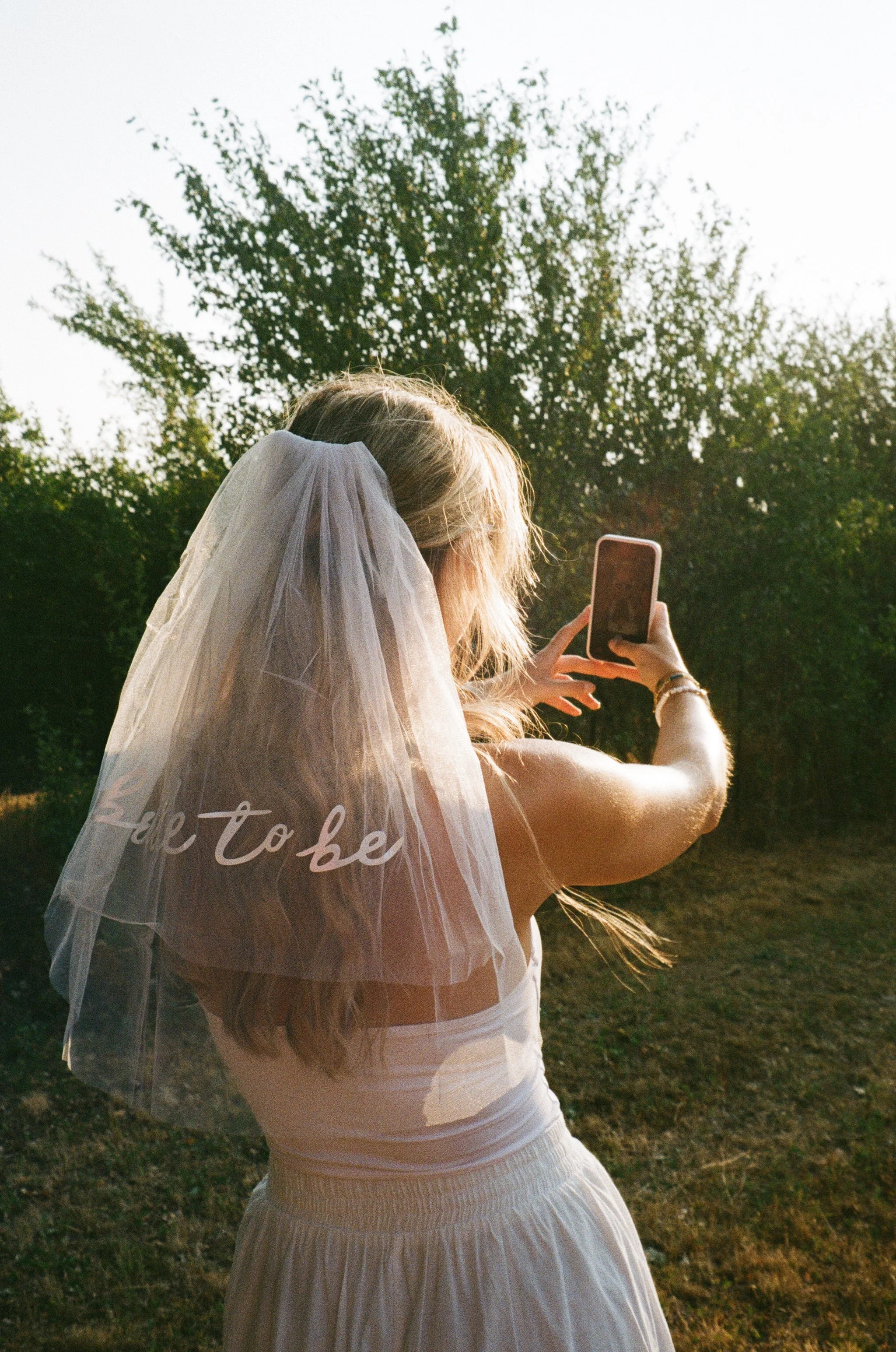A woman dressed in white with a veil takes a selfie outdoors with sunlight illuminating her hair, surrounded by trees and grass.