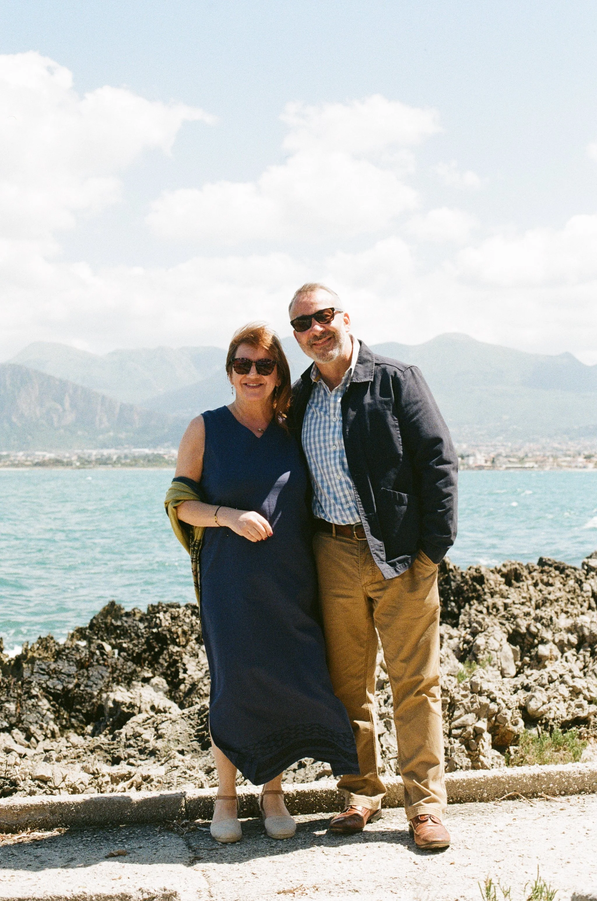 A happy couple standing on rocks by the water with a mountain range in the background, smiling and wearing sunglasses.