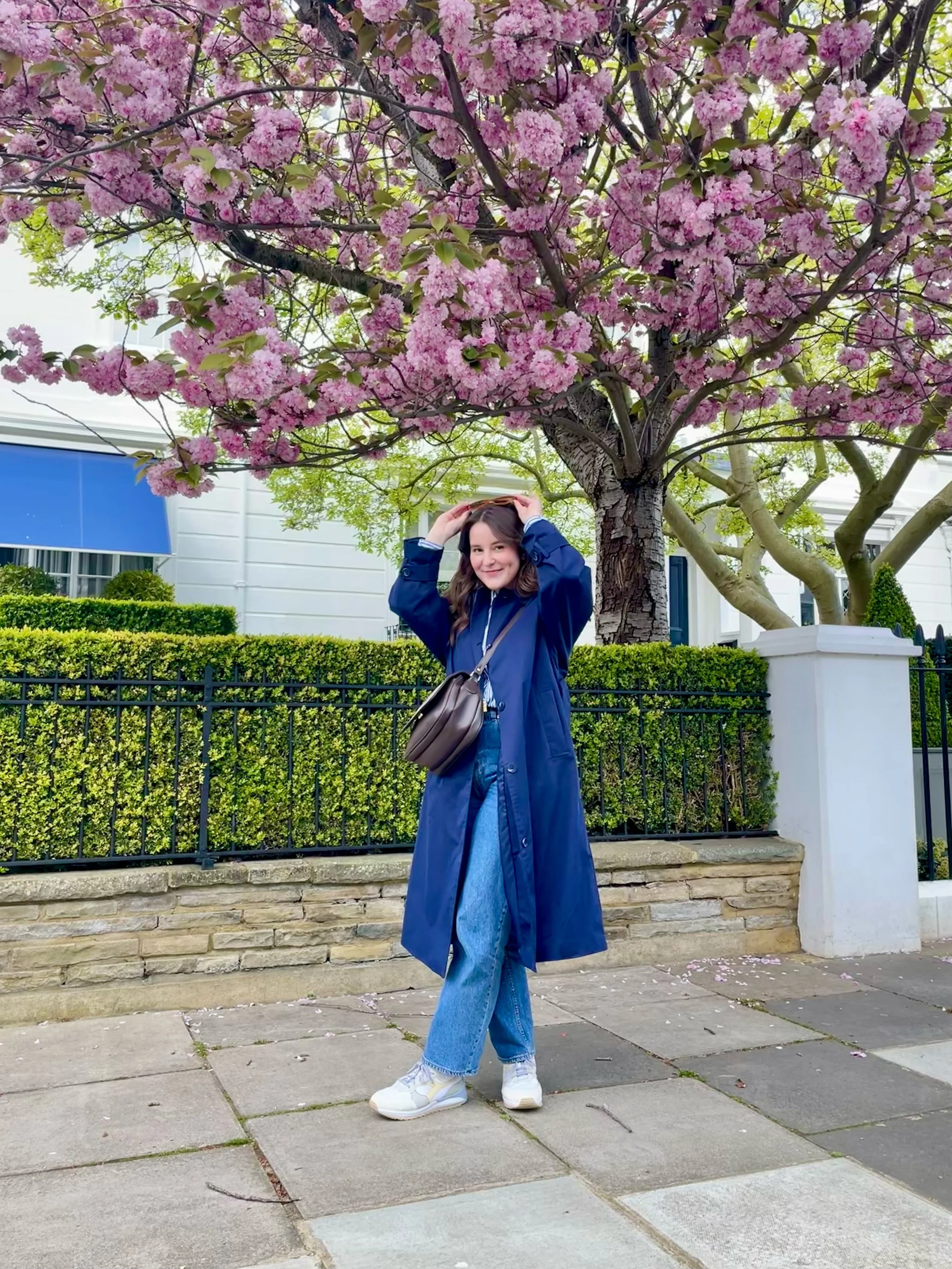 A young woman standing on a sidewalk in front of a black fence and a hedge, with a large pink flowering tree behind her. She is smiling and adjusting her hair, dressed in a blue raincoat, jeans, and white sneakers, carrying a brown purse.