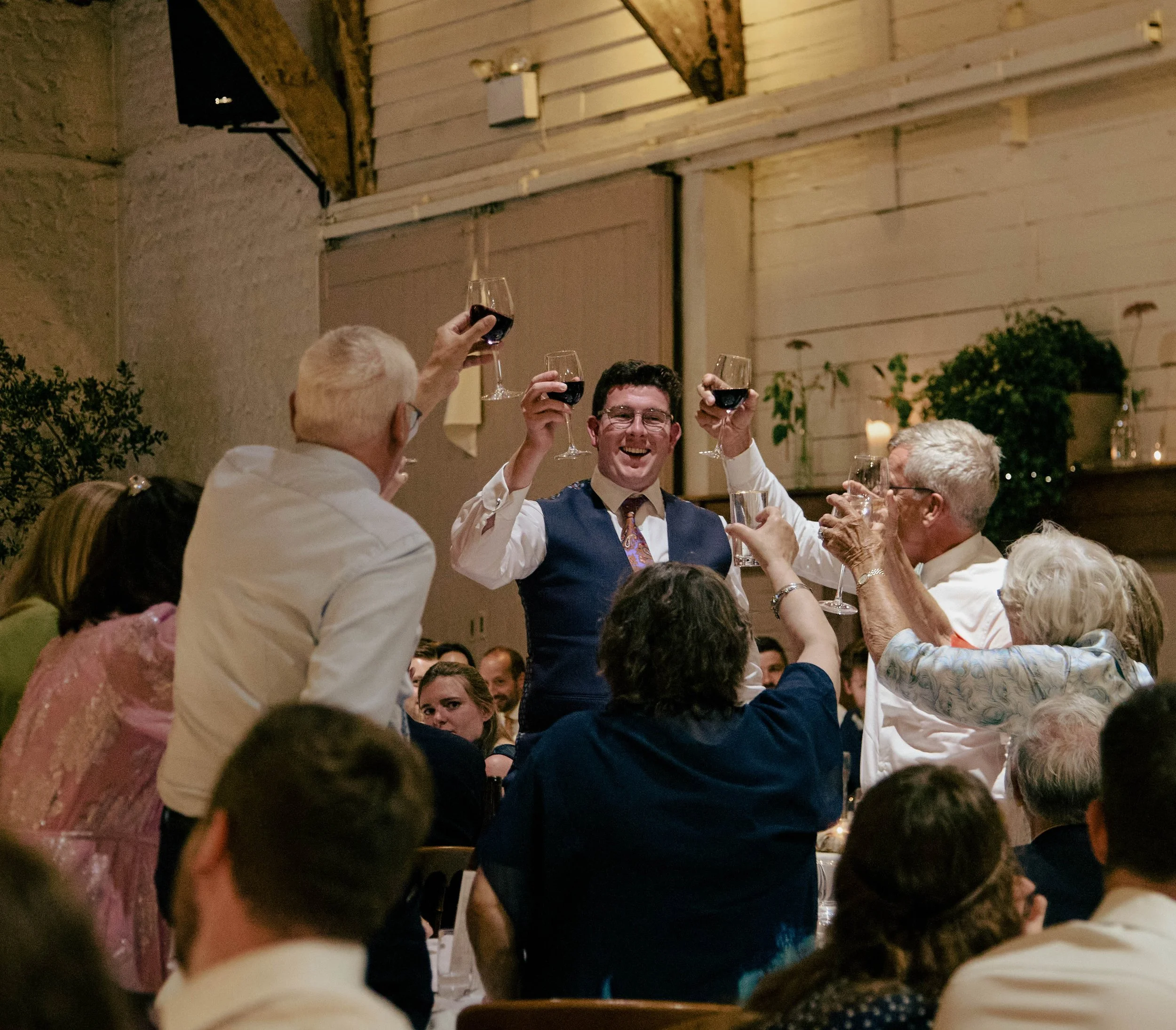 Group of people celebrating with glasses of red wine raised in a toast at a social gathering in a warmly lit room with a rustic interior.