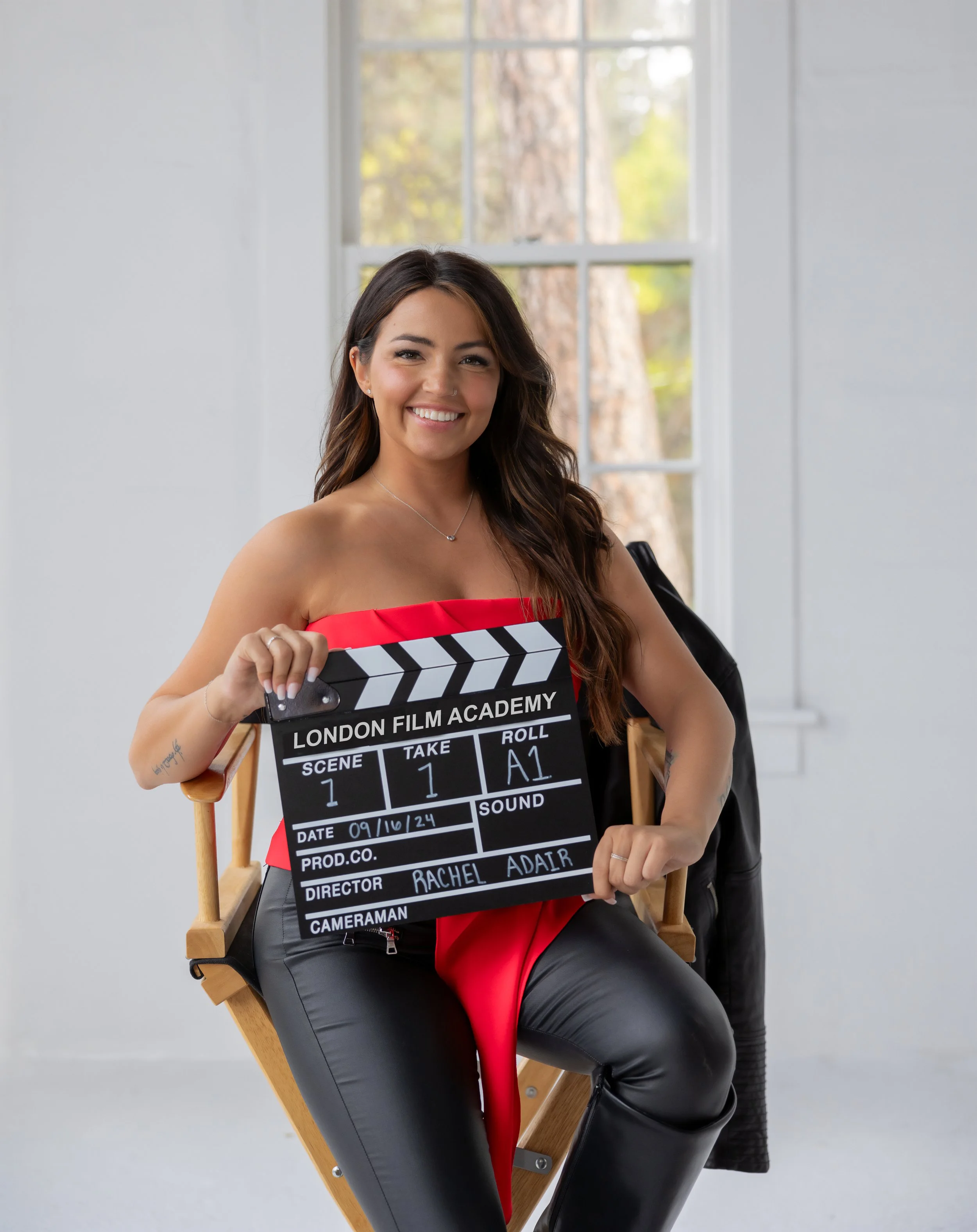 A young woman with long dark hair, smiling, sitting on a director's chair indoors, holding a film slate with 'London Film Academy' written on it, wearing a red top and black leather pants.