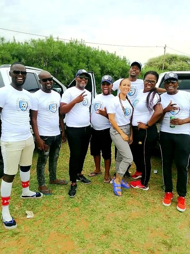 Group of eight people standing outdoors on grass, smiling and wearing matching white T-shirts with a blue graphic.