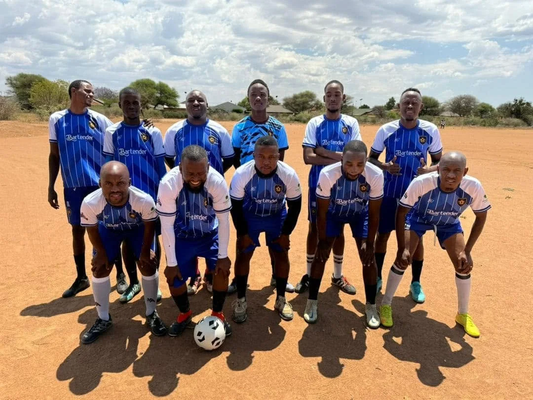 A group of eleven men on a dirt football field wearing blue and white soccer uniforms, posing for a team photo with some standing and some crouching, with a soccer ball in front of the crouching players. The background has a few trees and a cloudy sky.