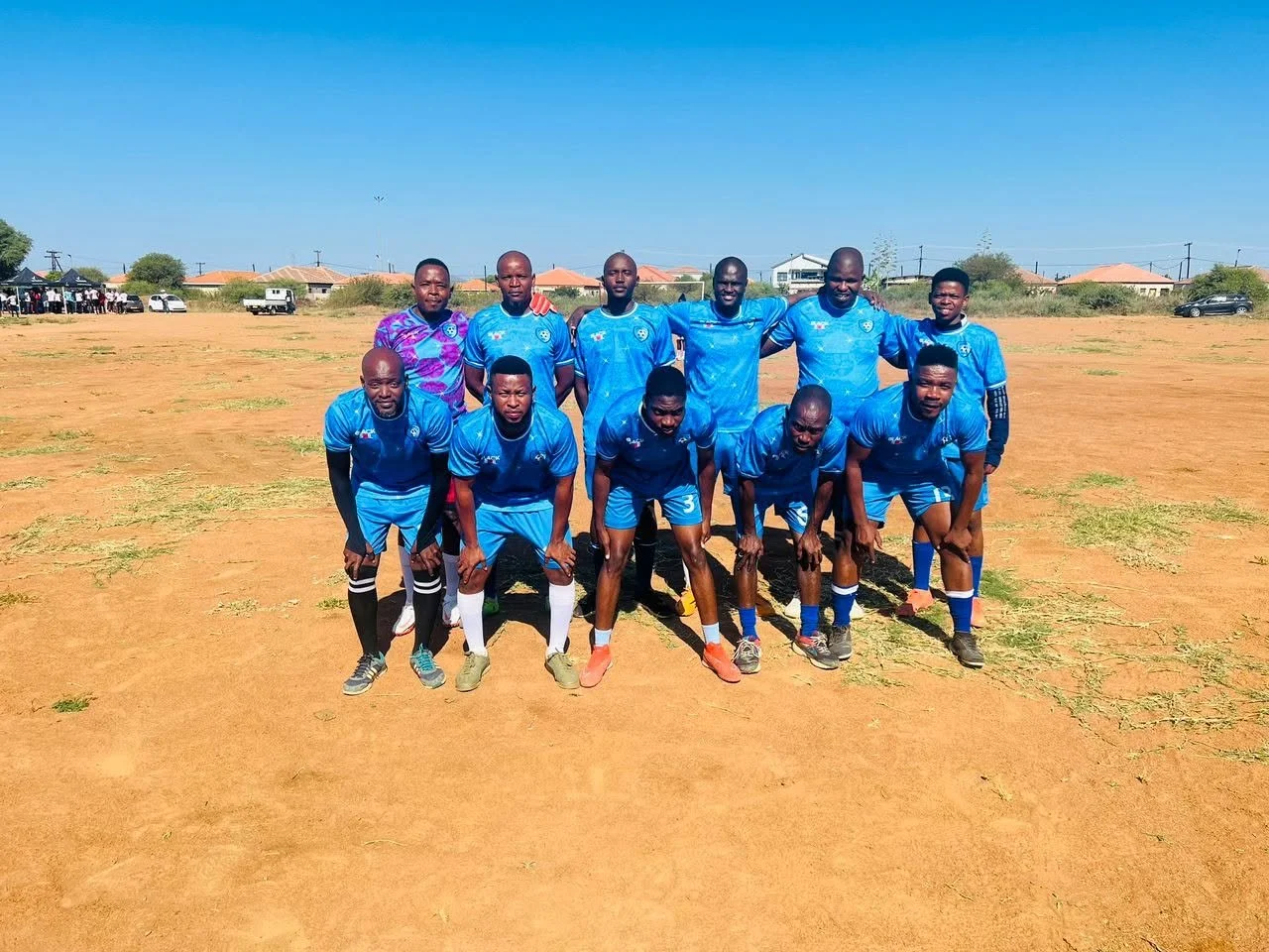 A group of eleven men in blue soccer uniforms posing on a dirt field, with some standing and others crouching, in a sunny outdoor setting with houses and cars in the background.